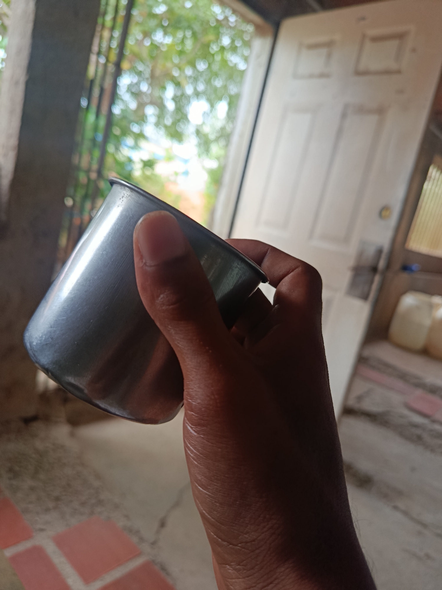 A hand is holding a silver metallic cup. The cup is partially visible and appears to be made of stainless steel. The background shows an interior space with an open white door, suggesting a domestic or casual setting. The environment is somewhat out of focus, but hints of foliage and a tiled floor are discernible. The lighting suggests it is daytime. The location is identified as Juan Griego, Venezuela.