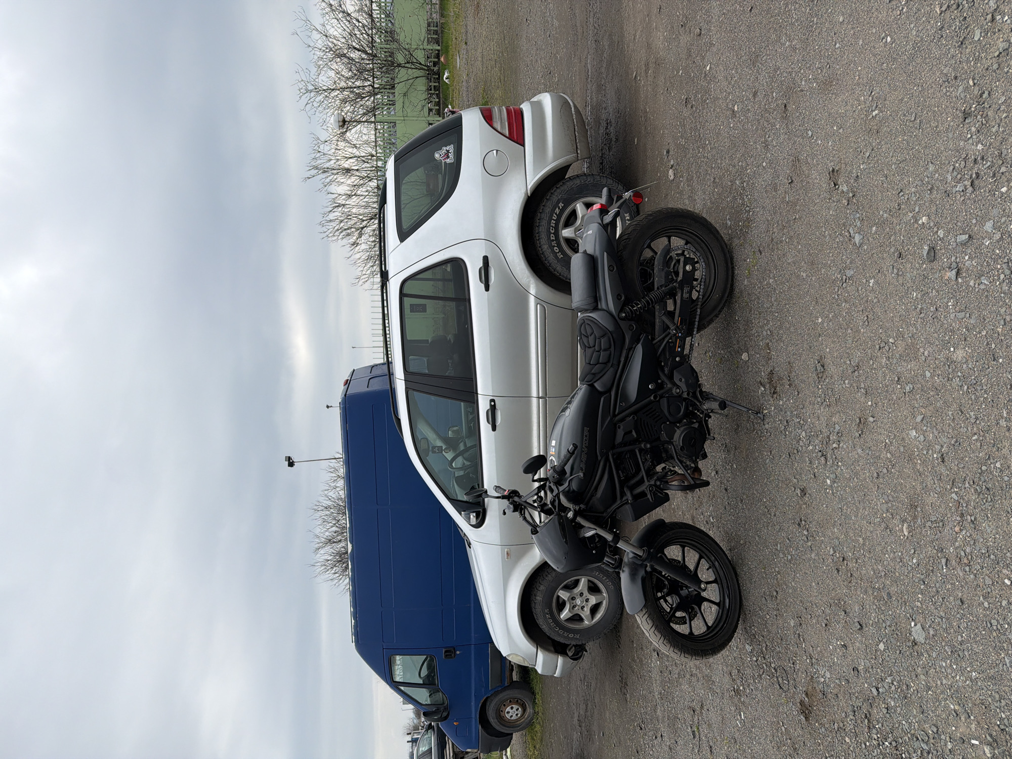 A black motorcycle is parked on gravel beside a white SUV and a blue van. The location is an open area, possibly a parking lot, with a sparse tree line in the background. The sky is overcast. The city of Burgas, Bulgaria is not verifiable from the image.