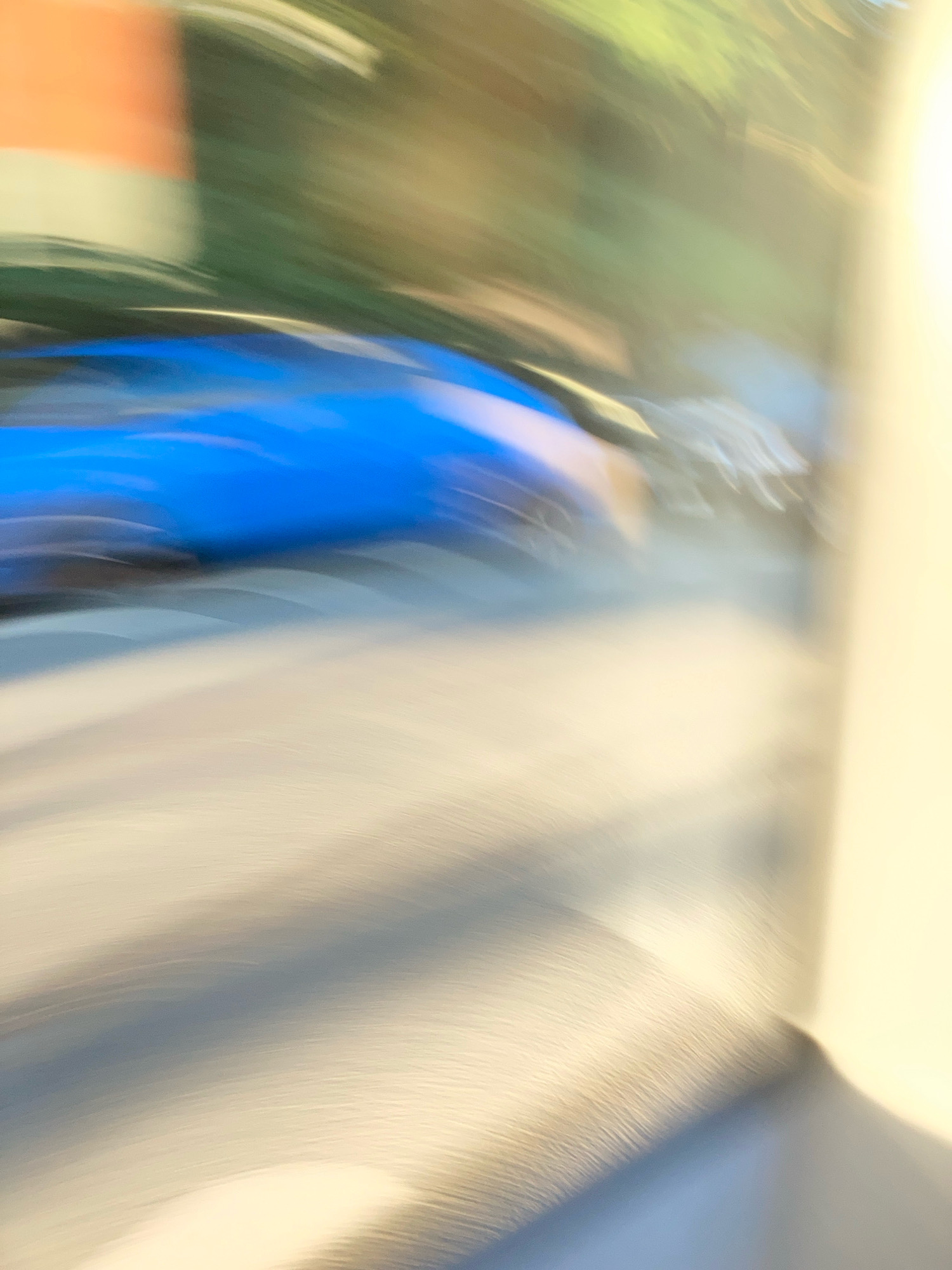 The image is an abstract, motion-blurred photograph. The primary subject appears to be a bright blue object, possibly part of a vehicle, with some lighter-colored shapes and streaks of green and orange in the background. The overall impression is one of movement and possibly an outdoor environment. There are no discernible people or specific landmarks that would allow for identification of a location in Prague, Czechia. The image is too abstract and lacks detail to provide a forensic or matter-of-fact description of a specific scene.