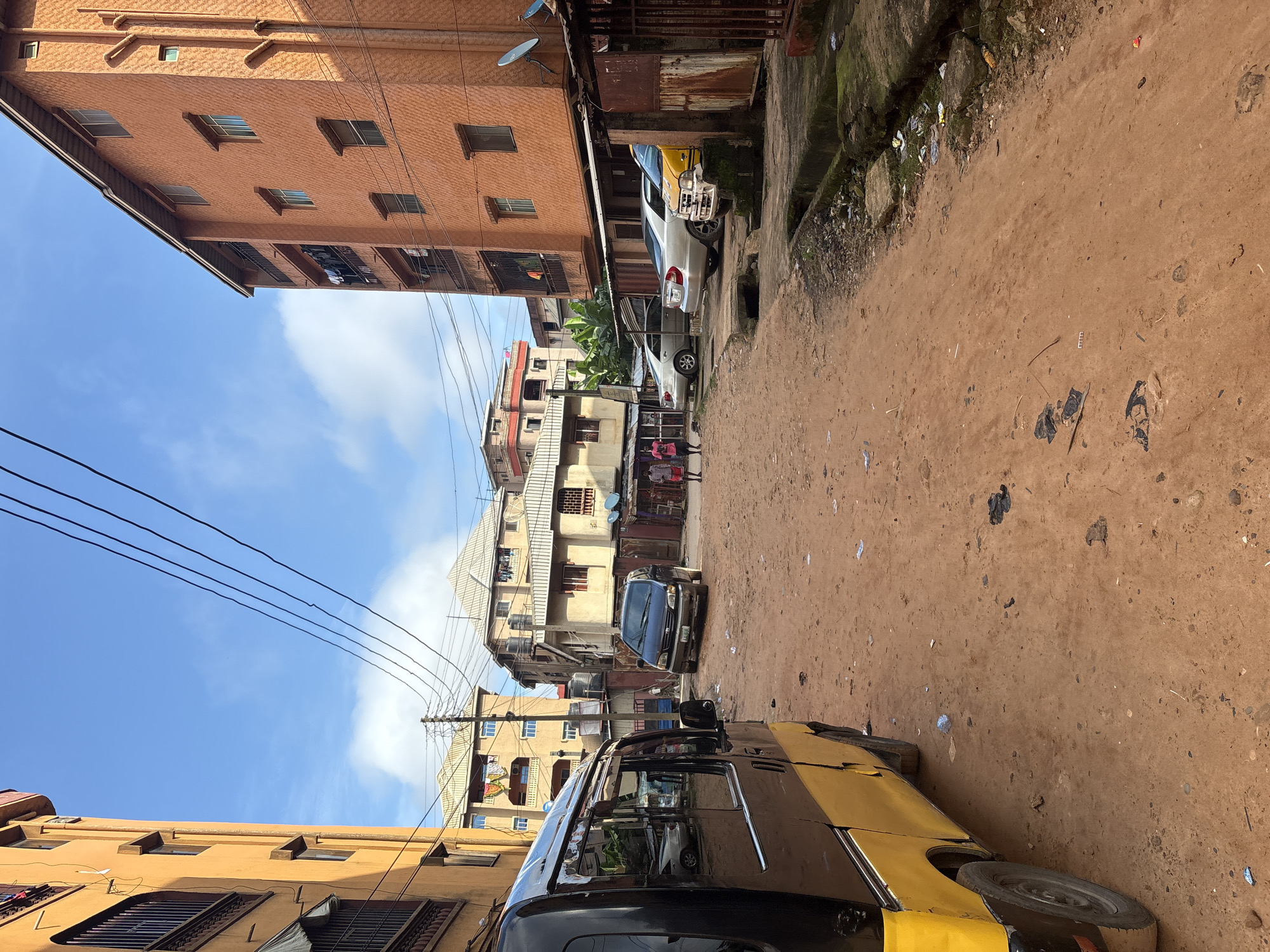 This is a daytime, eye-level, medium shot of a dusty street in Onitsha, Nigeria. The street is unpaved and appears to be made of red earth. There is trash scattered on the ground. The street is lined with multi-story buildings, typical of urban areas in Nigeria. On the left side of the image, a yellow and black van is parked. In the center of the street, there are several cars parked. Two individuals, appearing to be male, are walking in the distance, one on the left and another on the right. On the right side of the street, there is a building with orange-colored walls and balconies where laundry is hanging. Another building with lighter colored walls is visible further down the street. Power lines are visible crisscrossing the sky. The sky is blue with scattered white clouds.