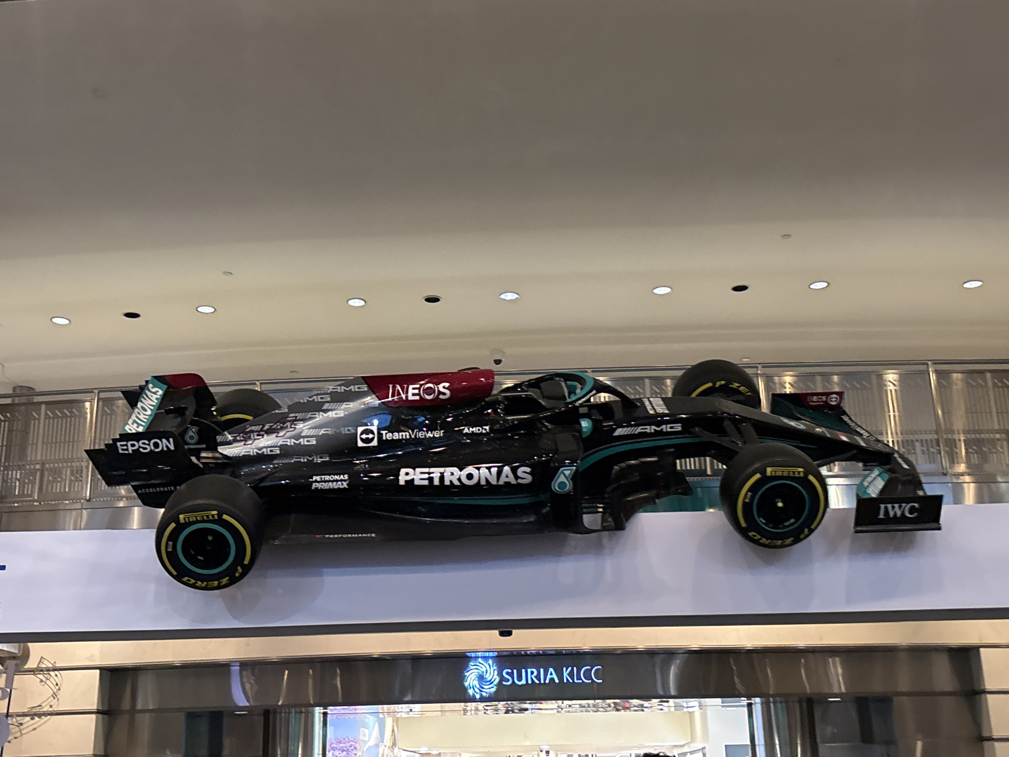 The image shows a black Formula 1 race car suspended from the ceiling of a shopping mall. The car is adorned with various sponsor logos, including PETRONAS, EPSON, INEOS, TeamViewer, AMD, and IWC. The location appears to be the Suria KLCC shopping mall in Kuala Lumpur, Malaysia, as indicated by the "SURIA KLCC" signage visible at the bottom of the image. The car is positioned horizontally and is viewed from a slightly elevated perspective, with the mall's interior and lighting visible in the background. There are no people visible in the immediate vicinity of the car.