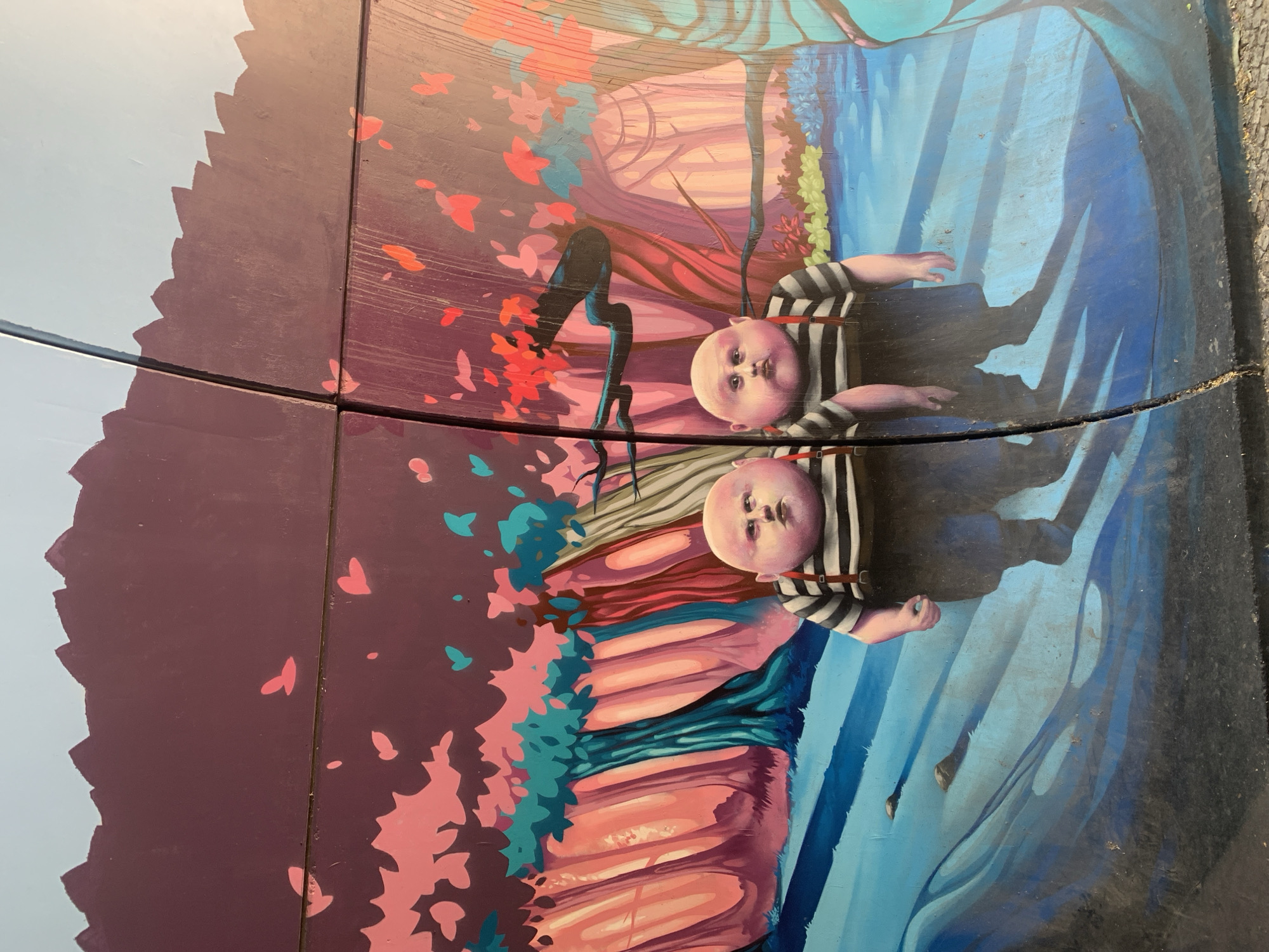 This image depicts a mural on a wall, likely part of an urban art project. The mural features two stylized, childlike figures with oversized heads and round bodies, wearing black and white striped shirts and dark overalls with red suspenders. They are positioned in a surreal, dreamlike forest setting with trees painted in shades of pink, purple, and turquoise. The ground appears to be a blue, watery landscape with dark shadows. The mural is divided by a vertical crack in the wall.
This mural is located on the exterior wall of the Pavilhão Multiusos de Odivelas in Odivelas, Portugal, as part of the "Festival MIRA" urban art festival. The festival transformed the exterior of the pavilion into a canvas for various artists. The figures in the mural are inspired by characters from "Alice in Wonderland," specifically Tweedledee and Tweedledum. The artwork aims to bring a touch of fantasy and artistic expression to the public space.