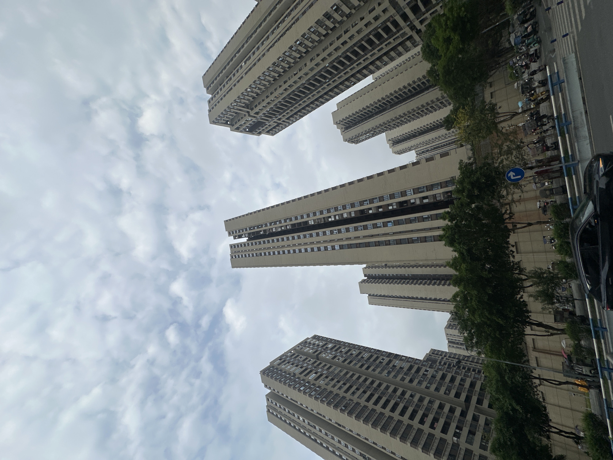 The image depicts a view of several tall, residential buildings in the Chongqing High-tech Zone, China. The perspective is from ground level, looking upwards. The buildings are constructed with a repeating pattern of windows and are a light beige color. A row of trees lines the right side of the frame, partially obscuring the base of the buildings. A street is visible on the right, with parked motorcycles and a traffic sign indicating a left turn. The sky is overcast with clouds. Part of a black vehicle is also visible on the right side of the frame.