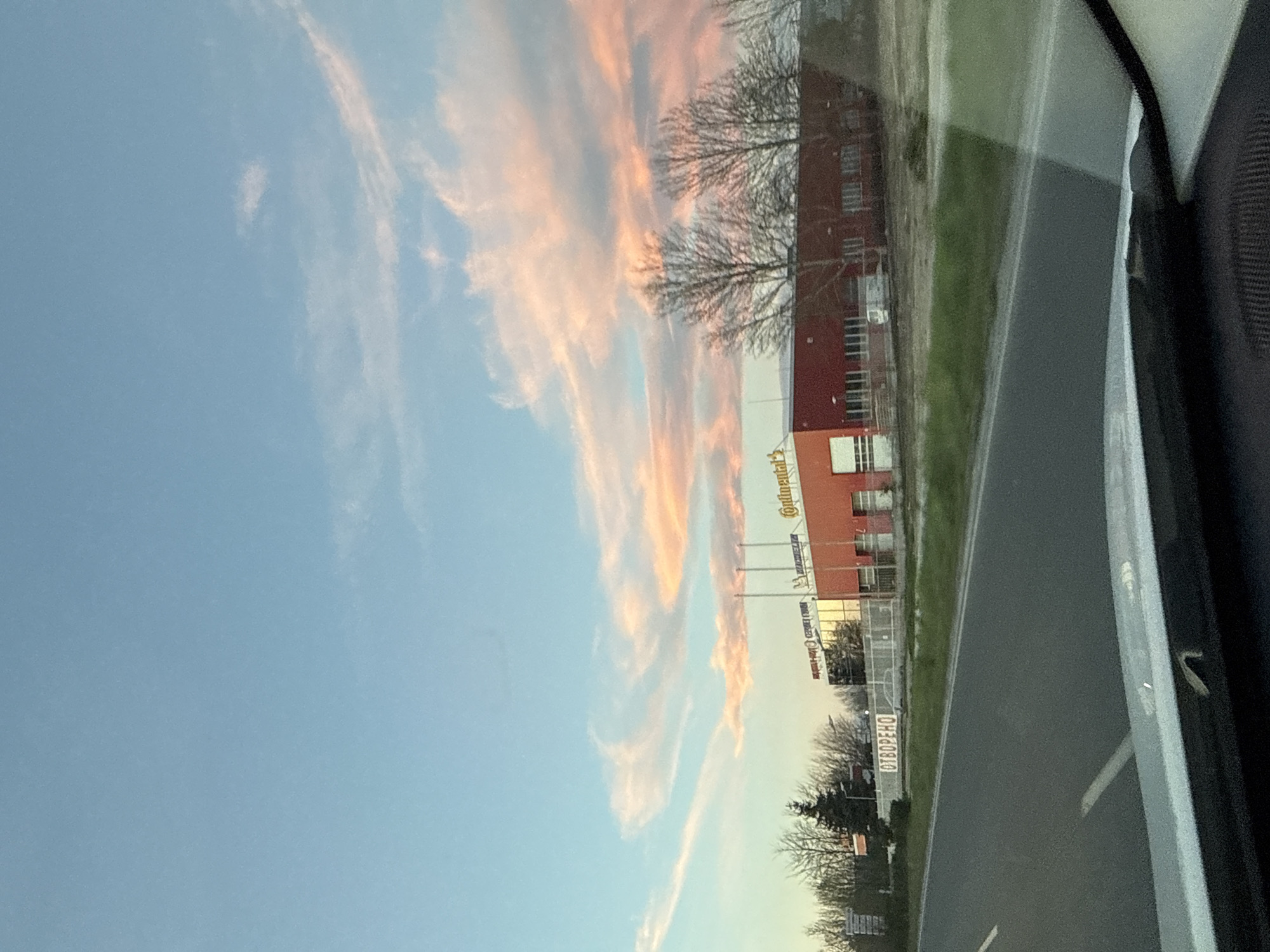 The image, taken from inside a vehicle traveling on a paved road, shows the sky, a building, and roadside details. The sky displays blue hues transitioning to pink and orange in the upper portion, suggesting either sunrise or sunset. The building is primarily orange and red with visible branding of "Continental" and "OTBOPEHO" written in Bulgarian. Bare trees are visible in the foreground. The road is grey, with some visible markings. The interior of the vehicle is partially visible on the right side of the image. Based on the branding and architectural style, the location can be identified as near the Continental tire factory in Plovdiv, Bulgaria.