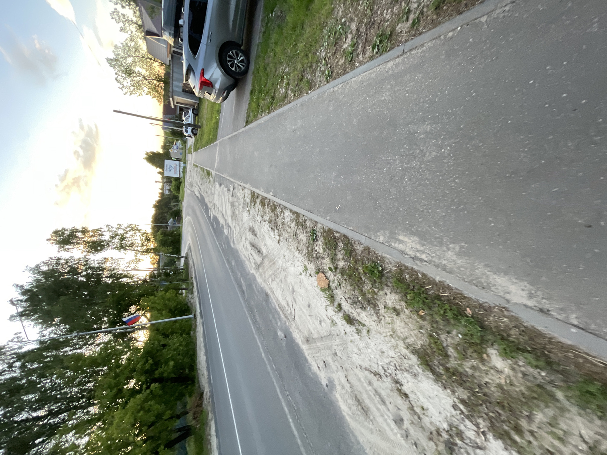 The image depicts a street scene in the village of Krasnye Baki, Russia, under a pale sky with scattered clouds, possibly at dusk. The perspective is from a sidewalk, looking down a paved road that curves to the left. To the left of the road is a sandy verge with tire tracks and some sparse vegetation. On the right side of the road, there is a paved sidewalk that runs parallel to the road. Beyond the sidewalk, there is a grassy area with some scattered debris and a few houses and garages. A silver car with a roof rack is parked on the right side of the road. In the distance, a Russian flag is visible on a flagpole, and there are trees lining the side of the road. The overall environment appears to be a semi-rural or suburban area.
