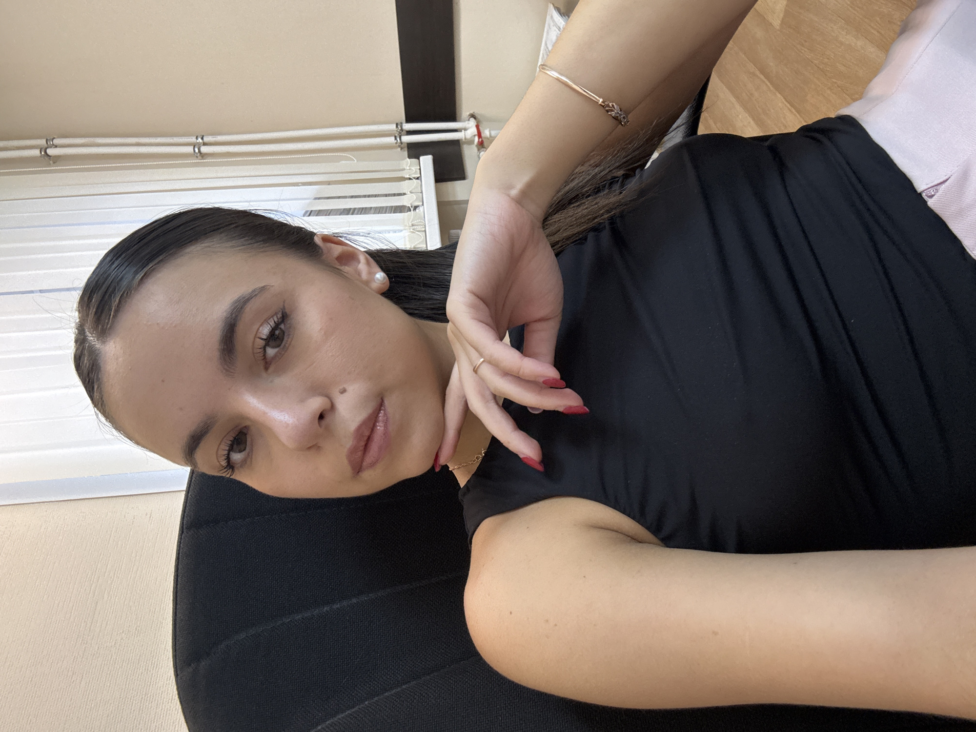 A young woman with dark hair pulled back is seated in a black office chair, facing the camera. She is wearing a black sleeveless top and a light pink skirt. Her left hand is resting on her chin, and she is wearing a ring on her right ring finger and a bracelet on her right wrist. She has on pearl earrings. The background shows a light-colored wall, vertical blinds, and a wooden floor. The overall lighting suggests an indoor, daytime setting.