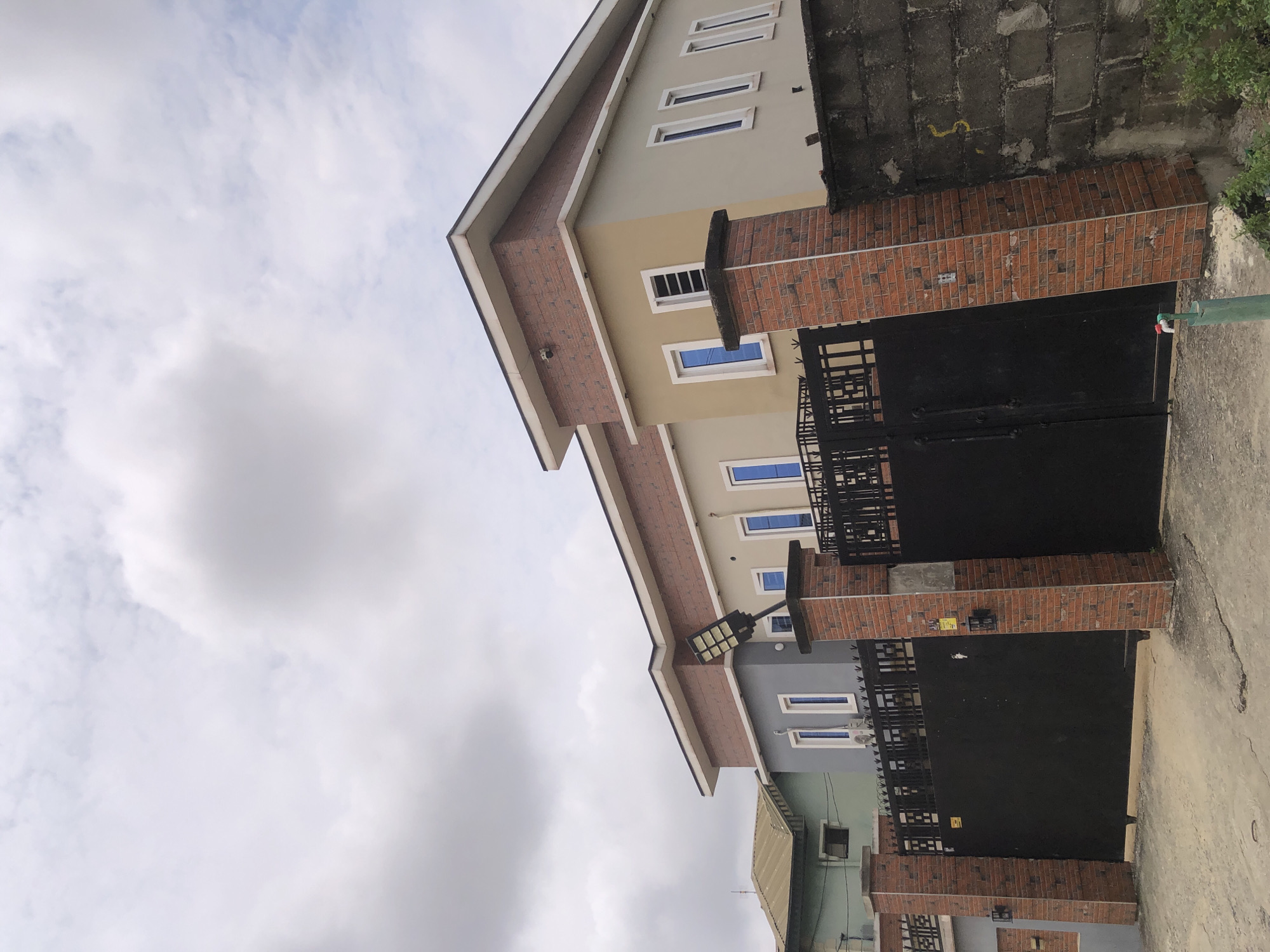 The image depicts the exterior of a modern residential property in Ado, Nigeria. The property features a multi-story house with a neutral facade, accented by brickwork on the upper levels and around the gate pillars. Large, dark metal gates provide access to the property, and are flanked by decorative brick pillars. A solar-powered street light is mounted on a brick pillar near the gate. In the background, other buildings are visible, including a house with a green facade and a corrugated iron roof. The sky is overcast with clouds. The ground in front of the gates appears to be paved. A green pipe with a faucet is visible in the lower right corner, suggesting an outdoor water source. The scene is static, with no people or immediate actions taking place.