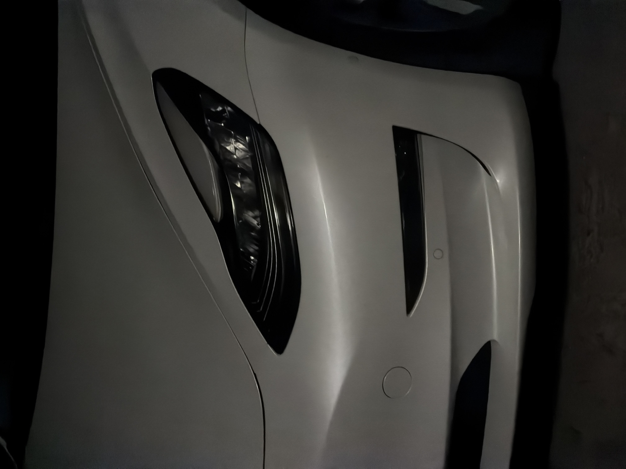 The image displays a close-up, low-angle shot of the front-right quarter of a white Tesla Model 3 car parked outdoors at night. The car's headlight and part of the bumper are visible. The environment is dark, suggesting it's nighttime. There are no people or animals in the frame, and no actions are taking place. The scene is a static observation of the vehicle. No specific location in Bila Tserkva, Ukraine, can be determined from the image.