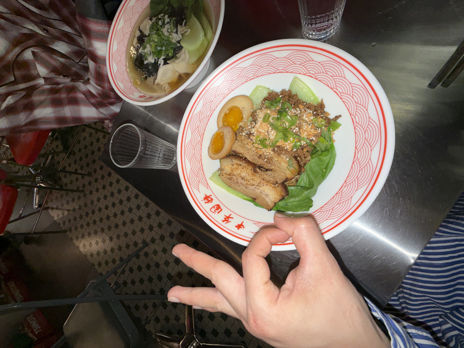 The image depicts a dining scene at Guo Xiang Niu Hua, a restaurant located in Paris, France. A light-skinned left hand, with a blue and white striped shirt cuff, occupies the left foreground, forming an "okay" gesture with its thumb and forefinger. On a stainless steel table surface, a white plate with a red scalloped rim and Chinese characters "牛華國祥" holds a meal comprising two slices of braised pork belly, a mound of minced meat garnished with chopped scallions and crushed peanuts or sesame seeds, two halves of a soft-boiled egg, and steamed bok choy. Adjacent to this plate, a white bowl with a matching red rim contains a clear broth noodle soup, featuring wontons, bok choy, seaweed, and chopped scallions. A clear, ribbed drinking glass stands between the two dishes. Another individual, partially visible in the upper right, wears a red and white plaid shirt. The background reveals a dark tiled floor, red bar stools with metallic frames, and obscured wall decorations, including visible text "TSINGTAO". A pair of chopsticks is partially visible at the bottom right of the frame.