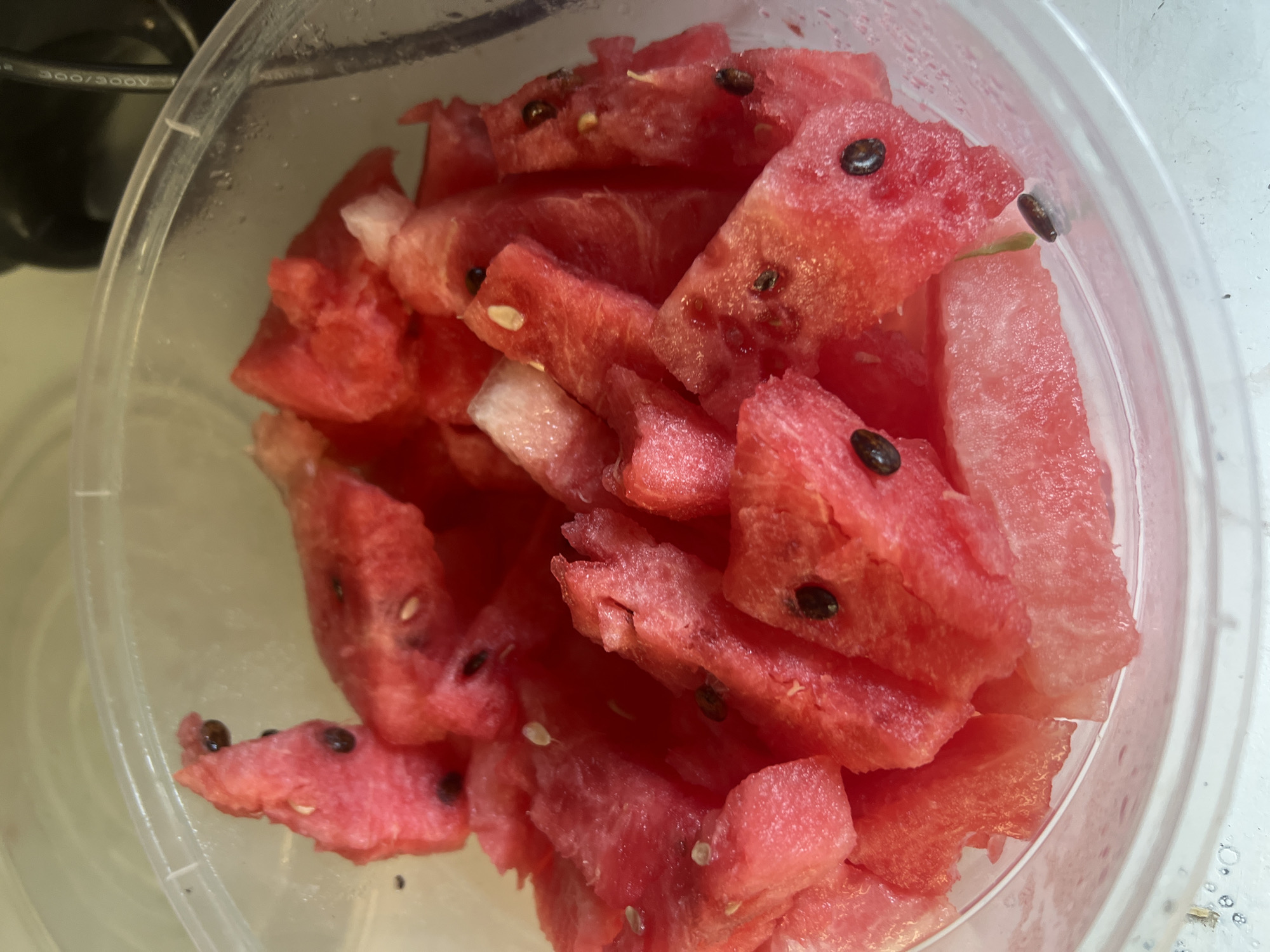 A close-up, overhead shot shows a clear plastic container filled with cut pieces of watermelon. The watermelon is a vibrant pink color, with some white rind visible on the edges of the pieces. Numerous dark brown watermelon seeds are scattered throughout the fruit. The container appears to be slightly damp on the inside, suggesting condensation. The background is blurred, but appears to be indoors, possibly a kitchen or refrigerator.
