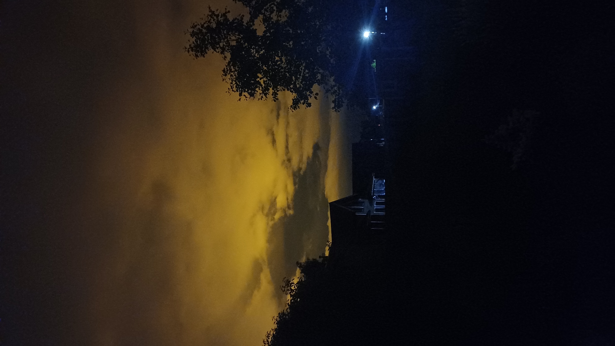 The image depicts a night scene, likely in Timokhovo, Russia, though specific contextual elements to definitively place the location are not visible. The sky is predominantly yellow-orange, likely due to light pollution illuminating low clouds or fog. A dark structure, possibly a building, is partially visible in silhouette against the sky on the right. Several light sources shine downwards. A tree silhouette is present, partially obscuring the view. The scene is generally dark, and the quality of the image is grainy.