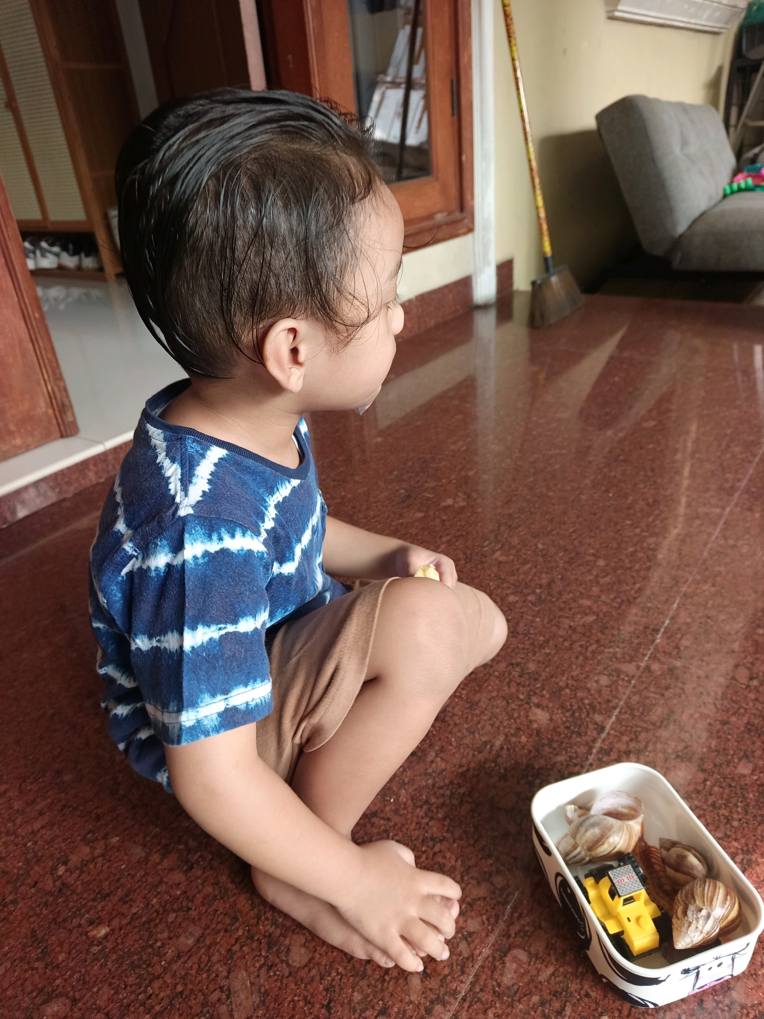 A young child with dark hair is sitting on a reddish-brown speckled tiled floor, facing to the right. The child is wearing a blue and white tie-dye t-shirt and light brown shorts. Their hands are resting on their knees, and their feet are bare. Next to the child is a white rectangular container filled with several seashells and a small yellow toy truck. The scene appears to be indoors, possibly in a home, with a wooden doorframe and a window visible in the background to the left. A broom with a long wooden handle and a grey cushioned chair are also visible in the background to the right. The lighting suggests it might be daytime. The child's expression is neutral, and they appear to be focused on something to their right. There is no visible text in the image. The setting suggests a casual, everyday moment at home in North Jakarta, Indonesia.