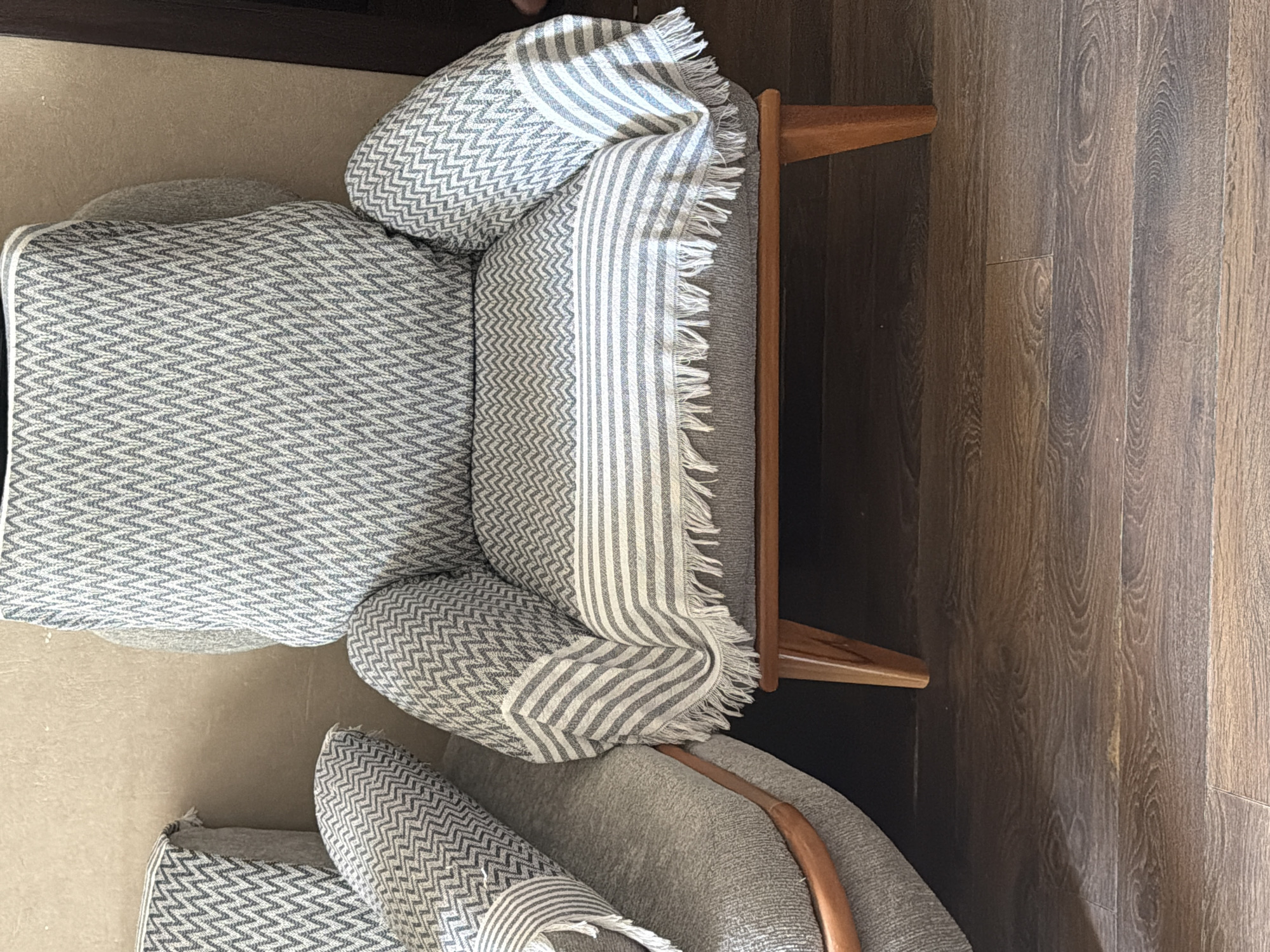 The image shows a close-up view of a grey armchair with a white and grey patterned throw blanket draped over it. The armchair has wooden legs and is placed on a dark wooden floor. To the left, a portion of another grey piece of furniture, possibly a sofa, is visible, also with a similar grey and white patterned throw. The background is a plain, light-colored wall. No people are present in the image. The scene appears to be an indoor domestic setting, possibly a living room. The location is identified as Tbilisi, Georgia.