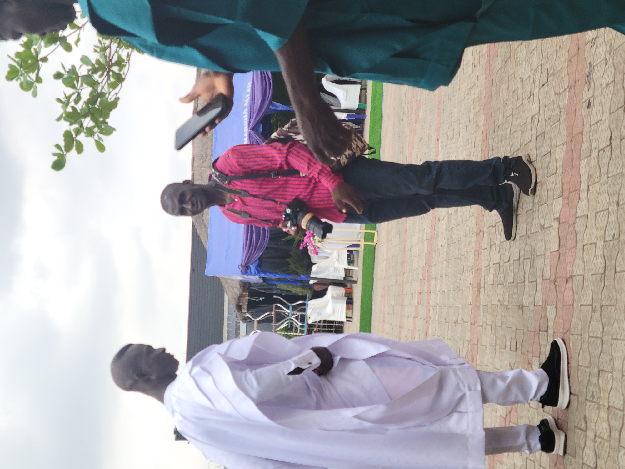 The image shows three men standing on a patterned paved surface. One man is wearing a long white robe and black shoes. A second man wears a red striped shirt, dark jeans, and black shoes. He has a camera strapped around his neck. A third man, partially visible, is wearing a teal shirt and holding a mobile phone. In the background, there is a blue and white draped structure with the words "MANSHA ALLAH" on it. White chairs and floral arrangements can be seen near the structure. The location is Suleja, Nigeria.
