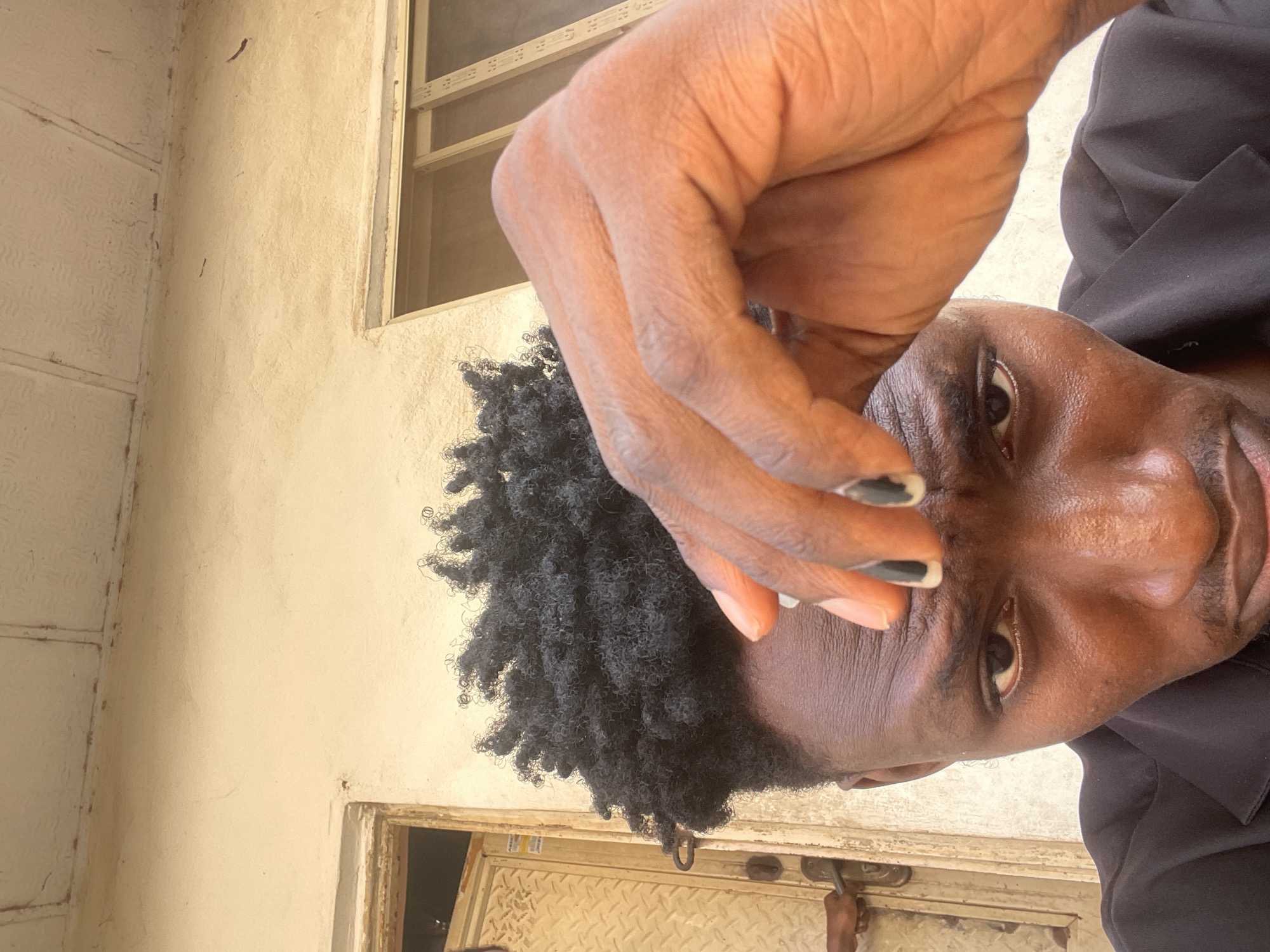A rotated portrait-oriented selfie shows a person with dark skin and short, kinky black hair, looking directly at the camera. The person is wearing a black collared shirt or jacket. The right hand is raised, with fingers partially obscuring the forehead and the upper part of the visible left eye. The visible fingernails (index, middle, and ring fingers) are painted black. The visible portion of the face shows an attentive gaze and a slight hint of a smile on the visible corner of the mouth.
The background consists primarily of a plain, light-colored wall, likely stucco or painted concrete, with some minor discoloration and texture. In the upper right quadrant of the image, a portion of a window with horizontal bars or slats is visible. Below the person's head and to the left, there's a glimpse of what appears to be a door or another structural element with a metallic, possibly diamond-plate-like texture, and a door handle or latch. The bright lighting suggests it is daytime. The overall setting appears to be an outdoor or semi-outdoor space, consistent with architectural styles found in locations like Lokoja, Nigeria. There is no visible text in the image.