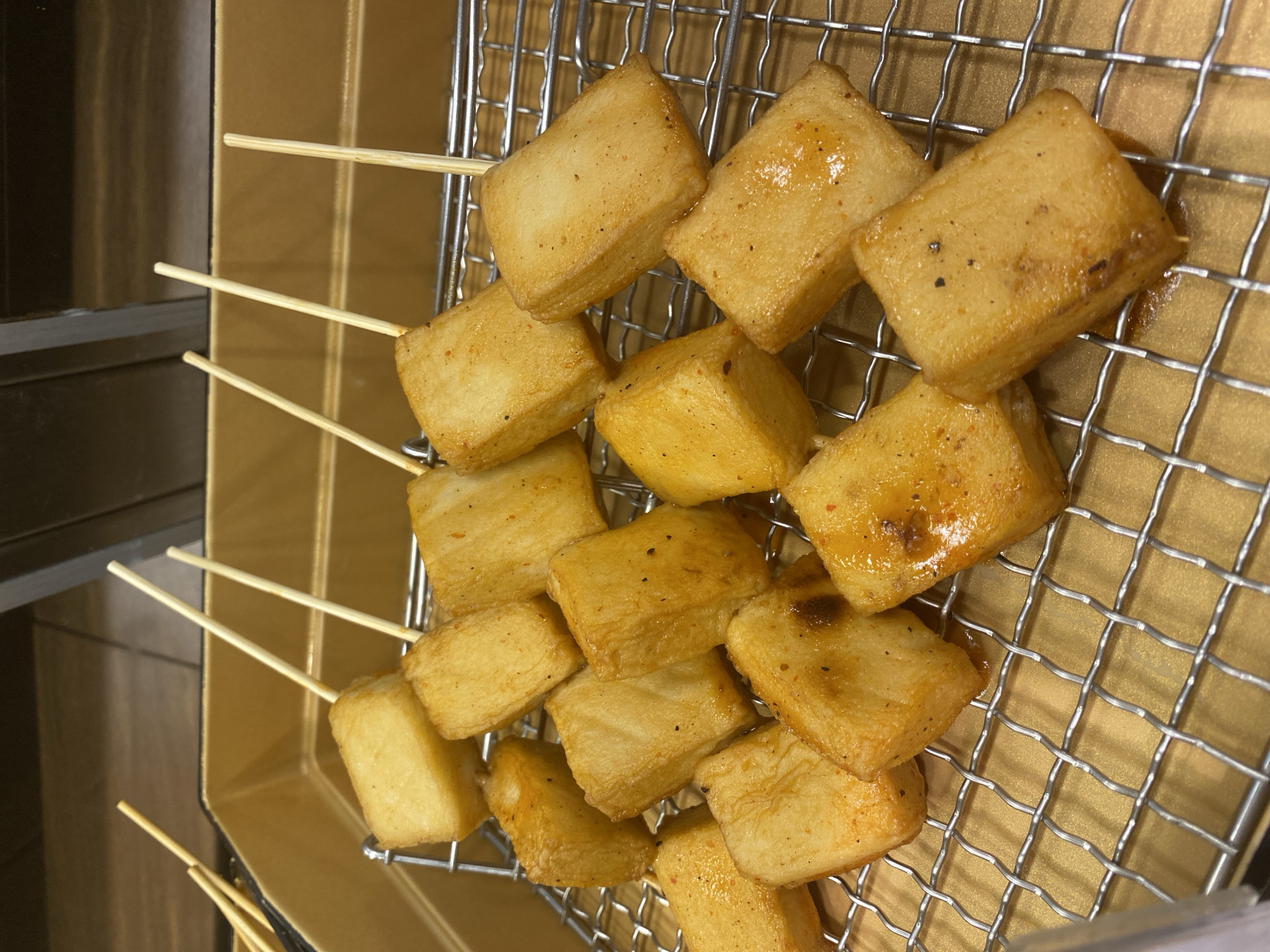The image shows a close-up view of several skewers of fried tofu, arranged on a metal wire rack over a golden-brown tray. The tofu pieces are cut into rectangular shapes and appear to be coated in a light orange seasoning, with visible specks of black pepper and red chili flakes. Some pieces show slight charring. The skewers are thin and made of wood. The background is out of focus but suggests an indoor setting with a metallic surface visible at the top. The lighting is bright, highlighting the texture and color of the food. The scene depicts food preparation or display, likely in a restaurant or market setting in East Jakarta, Indonesia.