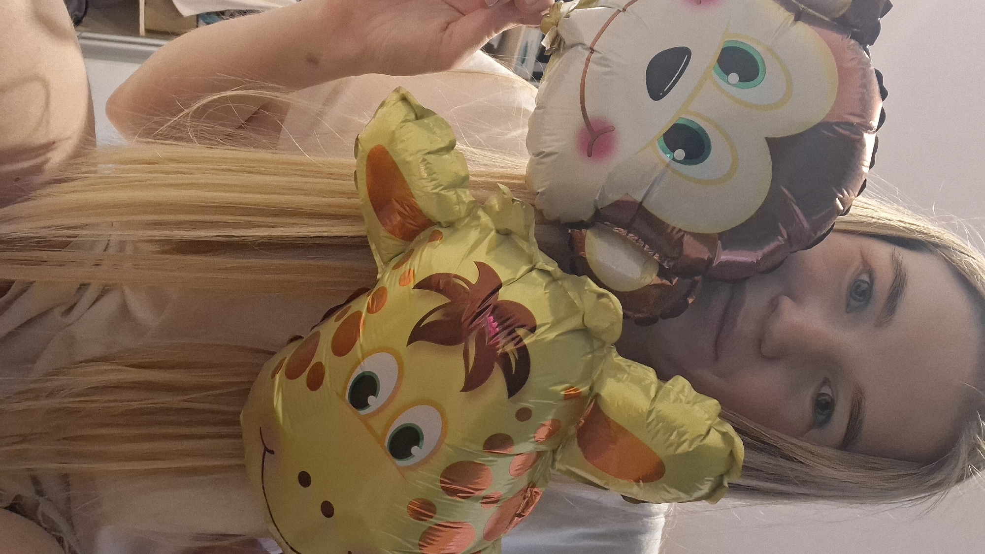A woman with light blonde hair is holding two inflated foil balloons. One balloon depicts a giraffe with brown spots, and the other is an owl. The background is a light-colored wall. The location is Chaikovsky, Russia.