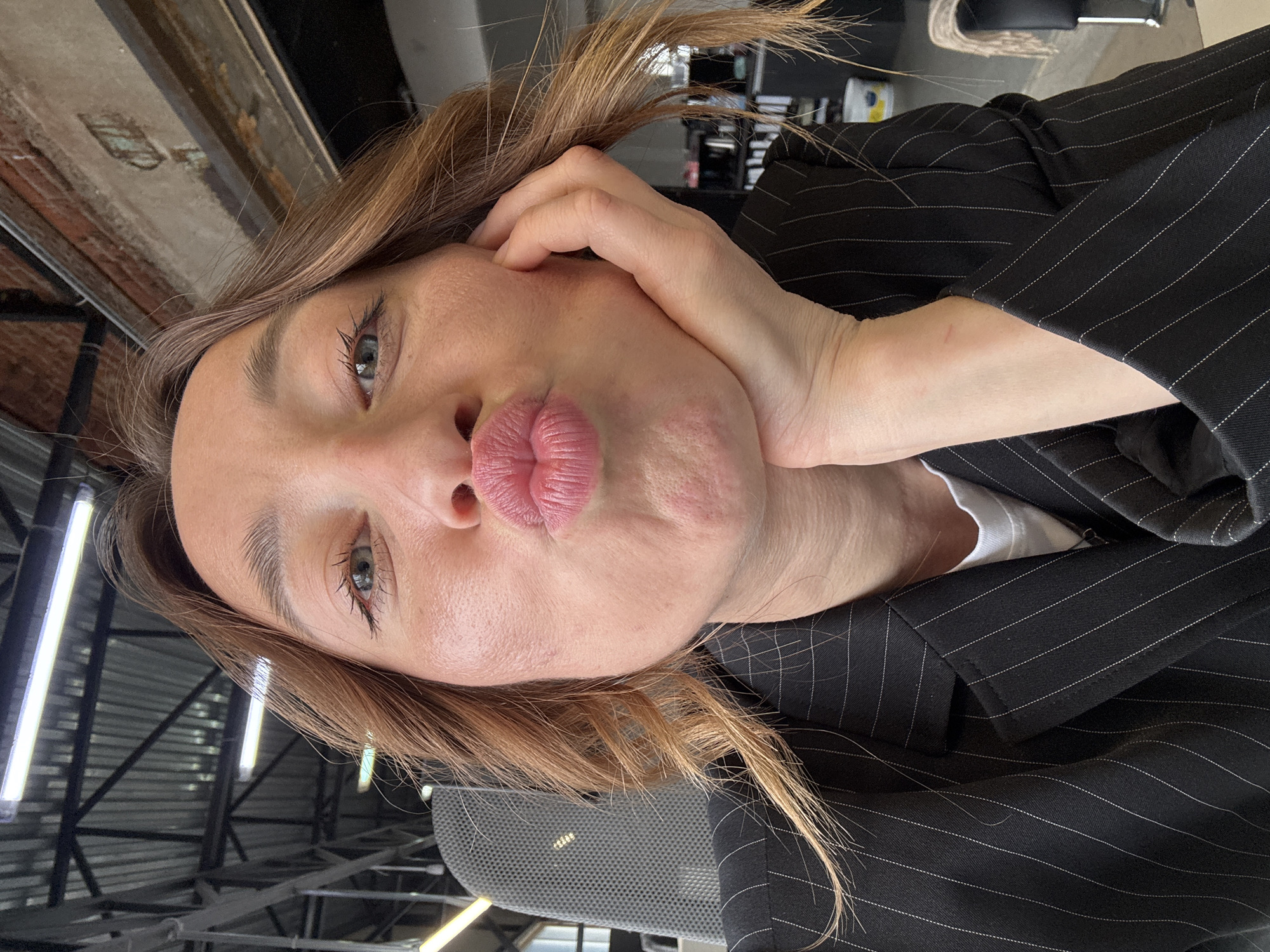 A medium close-up, eye-level shot features a young adult woman indoors, captured in a selfie-style portrait with the image rotated 90 degrees counter-clockwise. The woman, with light brown hair, occupies the central frame, making a playful "duck face" with her lips puckered. Her bright blue-green eyes are open and looking directly at the camera, complemented by well-defined eyebrows and visible eyelashes. Her right hand is gently cupping her chin and cheek.
She is dressed in a dark pinstripe blazer or jacket, worn over a white inner layer. The background suggests an industrial or contemporary office setting, characterized by exposed ceiling structures, a dark brick wall section, and several rectangular fluorescent light fixtures. Various elements like what appears to be a black chair or furniture and a mesh-patterned object are also discernible in the background. The lighting is artificial, emanating from the overhead fixtures. A small, faint red mark is visible on her right forearm. The scene takes place in Chaikovsky, Russia.