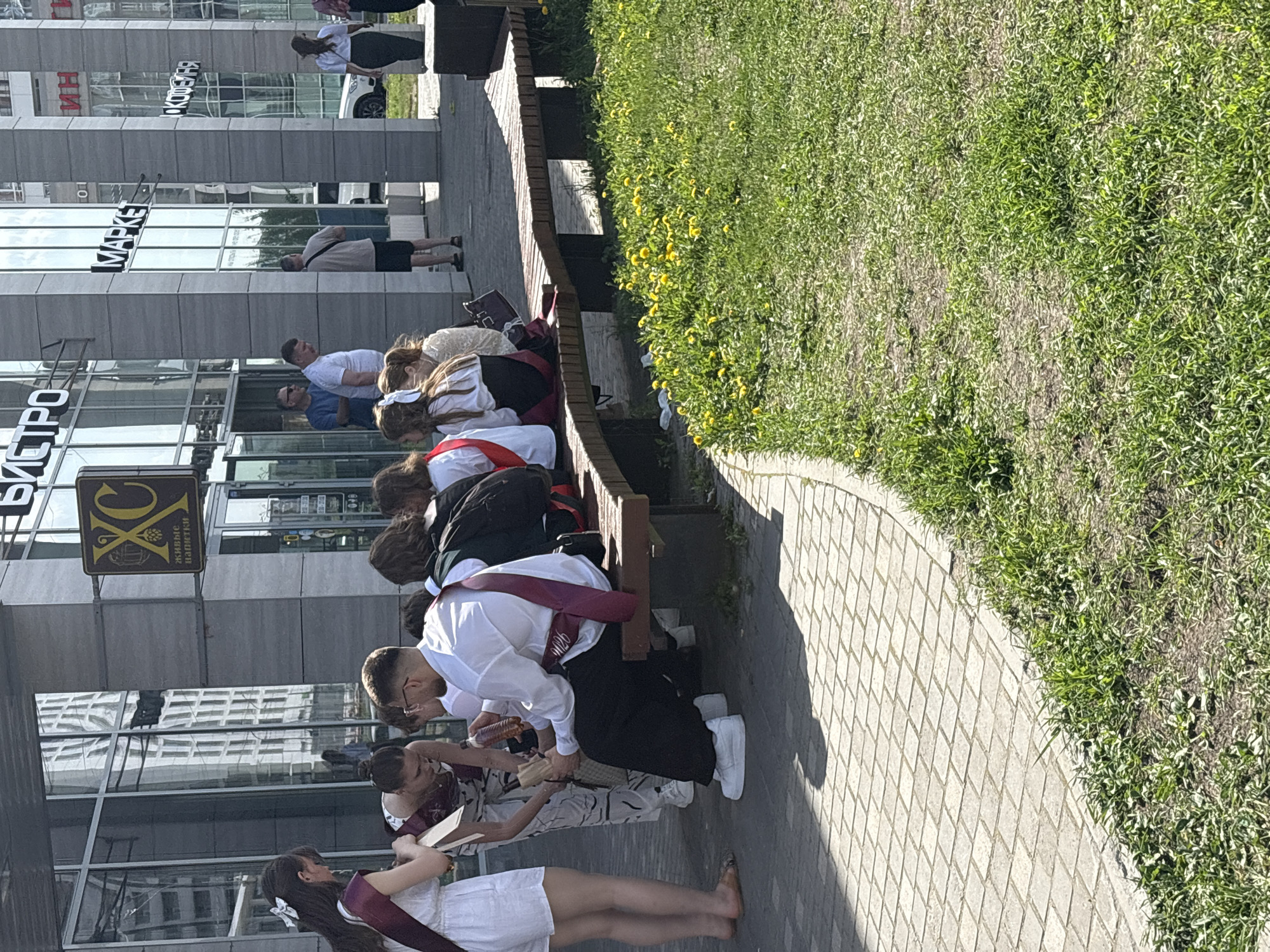 A group of students wearing graduation sashes pose for photographs along a city sidewalk in Perm, Russia. A bench lined with graduates is situated next to a walkway bordered by green space. The backdrop includes a modern glass-fronted building with visible signage reading "MARKET" and "БИСТРО." Other less legible signs adorn the building facade. Various pedestrians walk nearby. The weather appears sunny, casting shadows on the sidewalk.