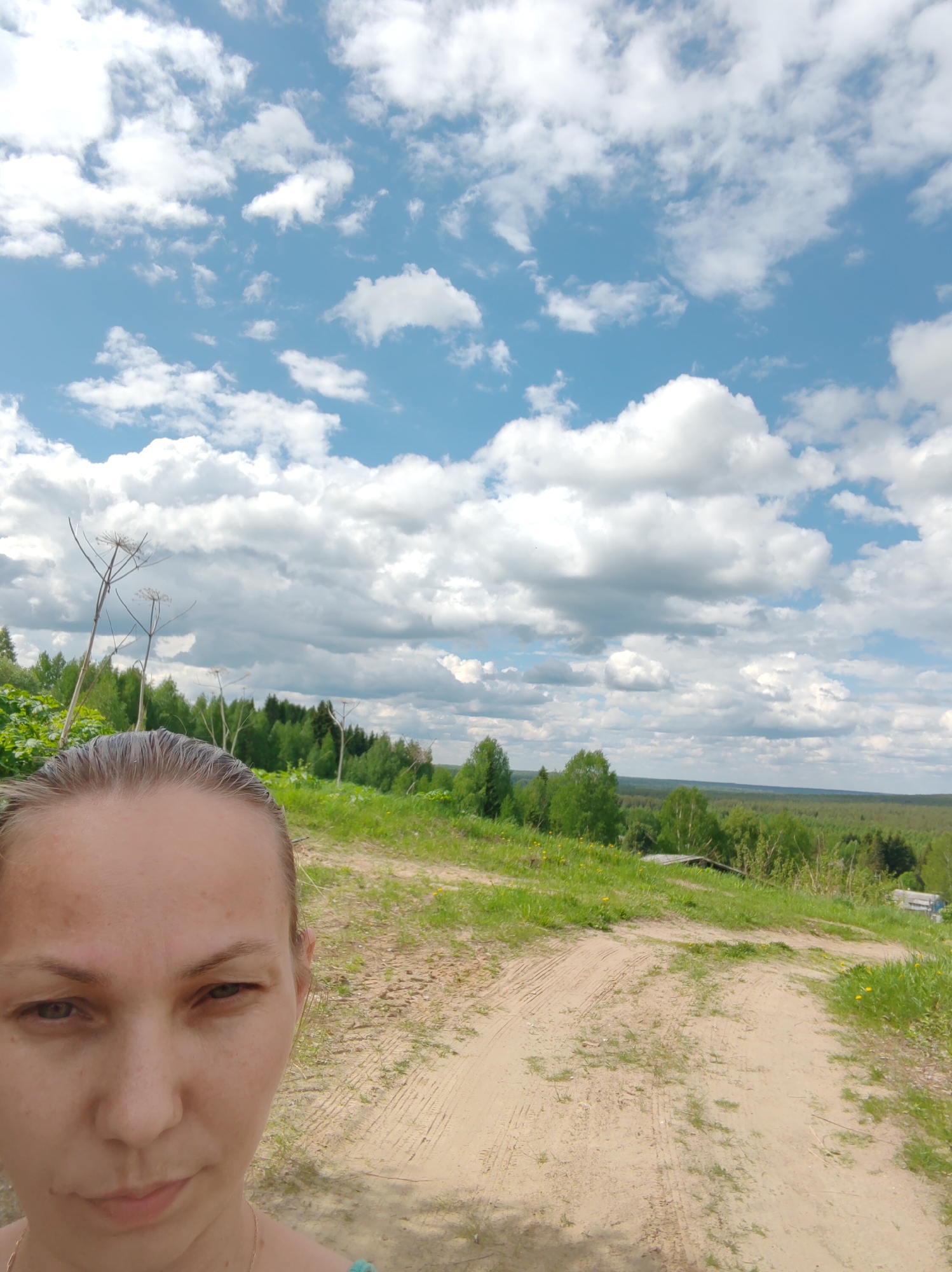 A selfie taken on a dirt road in a rural area. A woman is partially visible in the lower left corner of the frame. Behind her is a landscape featuring a grassy field, a dirt road with tire tracks, trees, and a distant forest. The sky is blue with scattered white clouds. The location is identified as the city of Чухлэм, Russia.