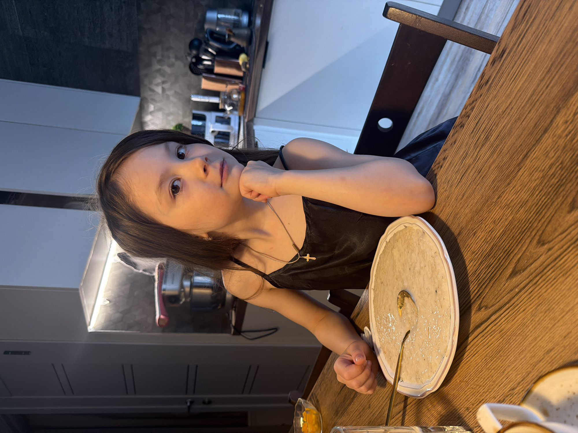 The image, taken in Perm, Russia, depicts a young girl sitting at a wooden table in a kitchen or dining area. The photograph is oriented vertically, with the scene rotated ninety degrees. The girl, who appears to be a child, has long, dark hair and looks directly at the camera with a thoughtful, slightly pensive expression. She wears a dark, possibly velvet or satin, camisole-style top and a delicate necklace with a small cross pendant. Her right hand is resting under her chin, while her left hand is positioned near a dish on the table.
In front of her, on the wooden tabletop, is a round, white plate containing a light-colored, somewhat thick or lumpy food substance, possibly porridge or a similar meal, with a spoon inserted into it. Partially visible at the bottom right is another plate or bowl. A small, clear glass or bowl with an orange substance, perhaps fruit or jam, is also on the table.
The background reveals a modern kitchen setup with dark-tiled walls and light-colored lower cabinets. Under-cabinet lighting illuminates the workspace, where various kitchen items like utensils, a grinder, and other appliances are arranged on the counter. A dark wooden chair back is also visible to the right. The lighting indicates an indoor setting, likely in the evening or under artificial light. No visible text is present in the image.