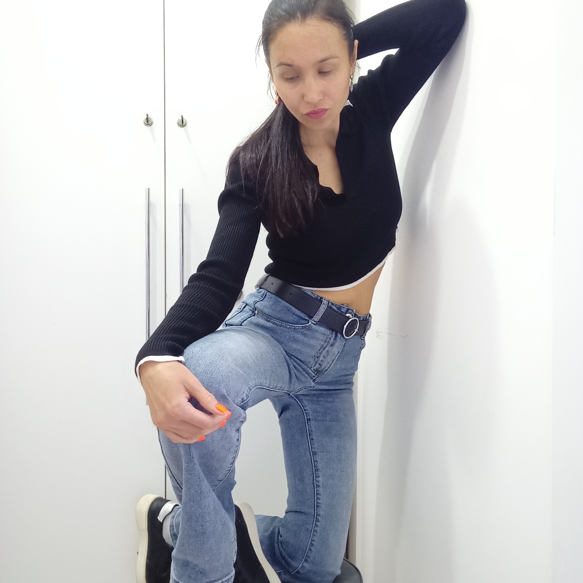 A woman is posing indoors, kneeling with her right leg on what appears to be a small, round seat or stool. She's wearing a black long-sleeved crop top with a white trim, blue jeans with a black belt, and black shoes with white soles. Her right arm is resting against a white wall, while her left hand is touching her right knee. She has dark hair pulled back into a ponytail and is looking down. The background consists of a white wall and a white closet or cabinet with silver handles. There is no indication that the image was taken in Izhevsk, Russia.