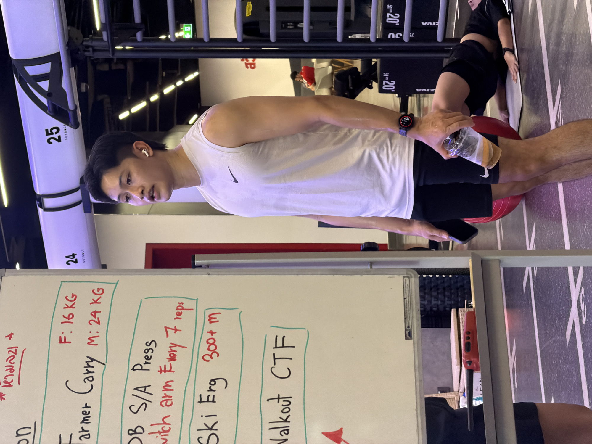 A young man in a white sleeveless shirt and black shorts stands in a gym in Nong Pa Khrang, Thailand, holding a clear plastic cup with a yellow beverage and a black smartphone. He wears white earbuds and a smartwatch on his left wrist. In the background, another person lies on a yoga mat with a red exercise ball behind them. A whiteboard lists exercises and weights, including "Farmer Carry" with "F: 16 KG" and "M: 24 KG," "DB S/A Press," "Ski Erg 300+ m," and "Walkout CTF." Gym equipment and lighting are visible.