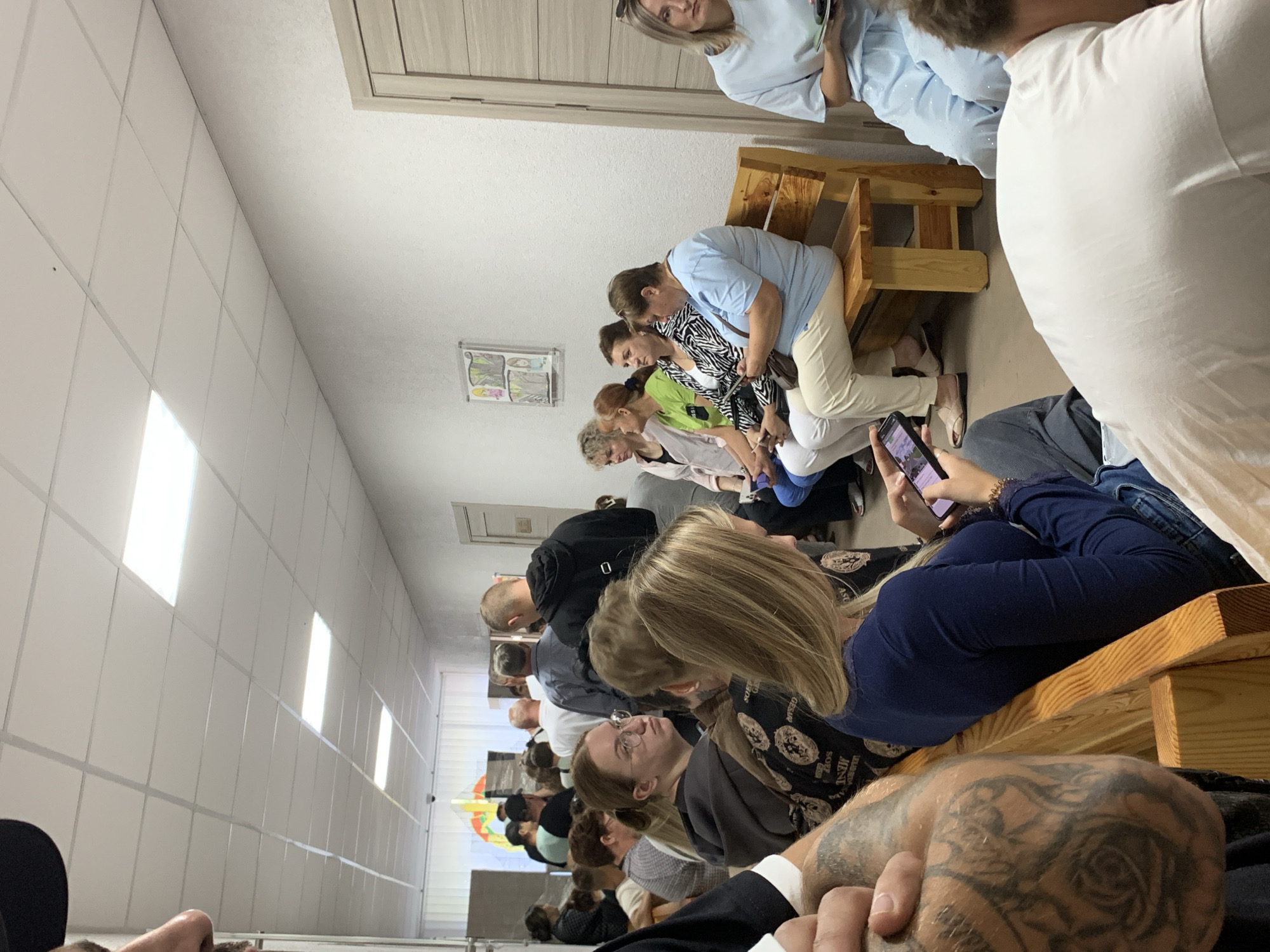 In a hallway with a tiled ceiling and white walls, a group of people are waiting. The hallway appears to be indoors, possibly a clinic or office. A row of fluorescent lights illuminates the space. On the right side of the hallway, there is a wooden bench where several individuals are seated. A woman in a light blue top and light-colored pants is seated on the bench, looking at a phone. Another woman with blonde hair in a dark blue top is seated in the foreground, also looking at a phone. A person with a tattooed arm is visible in the lower left of the frame. In the background, more people are standing and seated, suggesting a queue or waiting area. A door with horizontal wooden panels is visible on the far right. On the wall, a framed picture or poster hangs. The overall atmosphere is one of waiting and inactivity. The location is specified as Бокшыцкі сельскі Савет, Belarus.