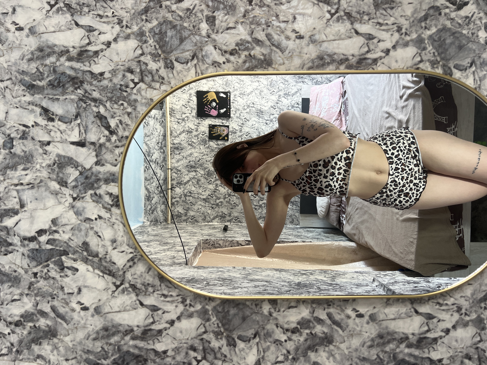 The image shows a young woman taking a mirror selfie in a bedroom. She is wearing a leopard-print pajama set consisting of a cropped top and shorts. The room has marble-patterned wallpaper and a bed with beige and pink bedding. There are two framed handprint artworks on the wall. The woman's tattoos are visible on her arm and leg. The mirror is oval-shaped with a gold frame and is positioned centrally in the frame. The overall scene suggests a casual, personal moment captured in a home environment.
