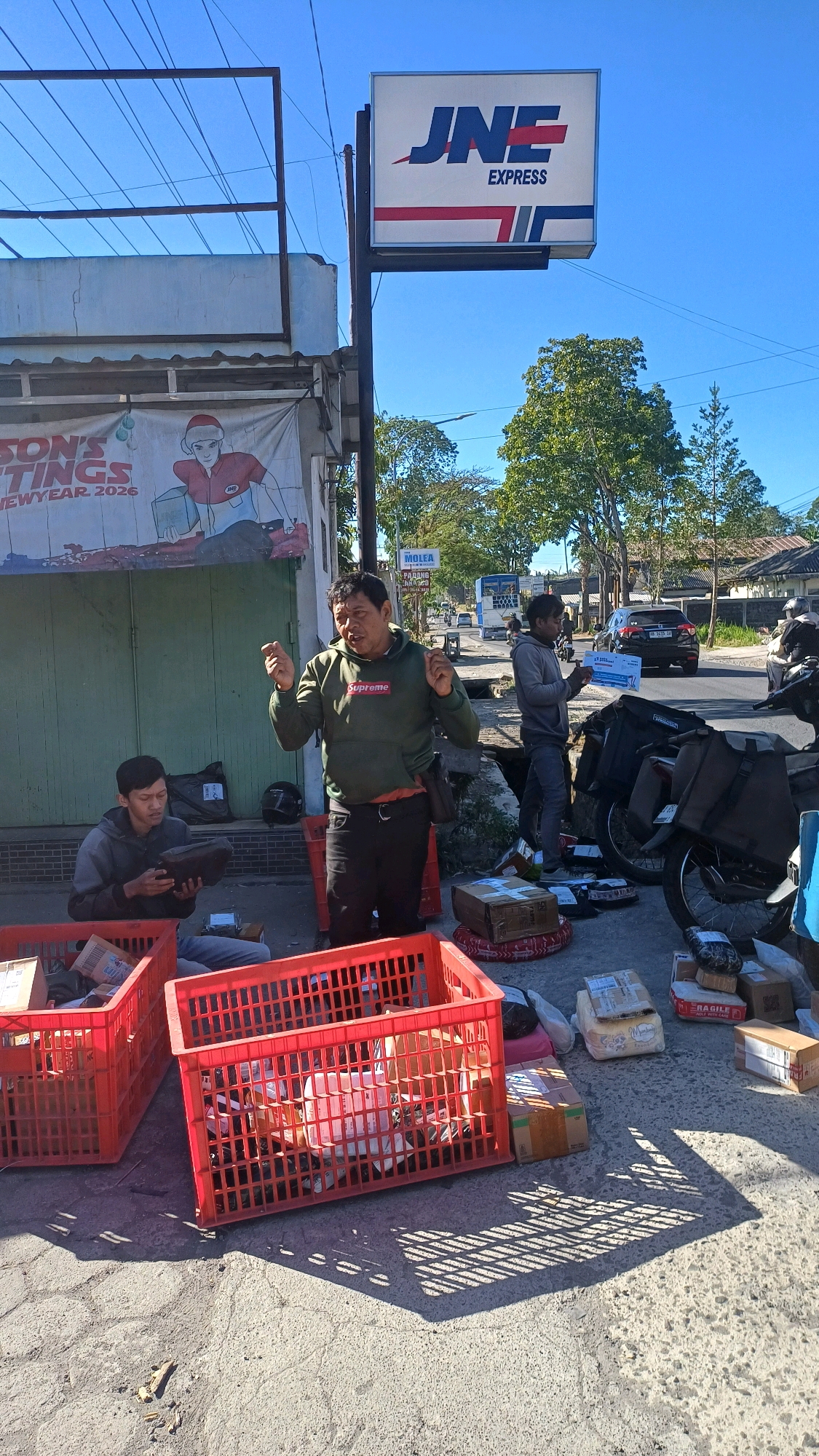 In Sosoran, Indonesia, on a sunny day, two men are engaged in a logistics operation outside a building with a prominent "JNE EXPRESS" sign. The building has a green door and a banner depicting a person in a Santa hat holding a package, with text that partially reads "SON'S TINGS NEWYEAR. 2026."
One man, wearing a green hoodie with "Supreme" written on it, is standing and gesturing with his hands as if explaining something. He has a dark belt and dark pants. Next to him, on the ground, are several packages and two large red plastic crates filled with more packages. A black helmet is also visible on the ground.
The second man is seated on the ground, wearing a grey hooded jacket and dark pants. He is holding a dark object, possibly a device or another package, and examining it. In front of him are also red plastic crates overflowing with packages.
Further in the background, another man in a grey hoodie stands by a motorcycle laden with bags. He is holding a document. A black car is visible on the road, along with other vehicles and buildings. The environment appears to be a roadside setting, with a pavement area for the men and their parcels, and a road for traffic. The scene suggests a busy delivery or sorting point.