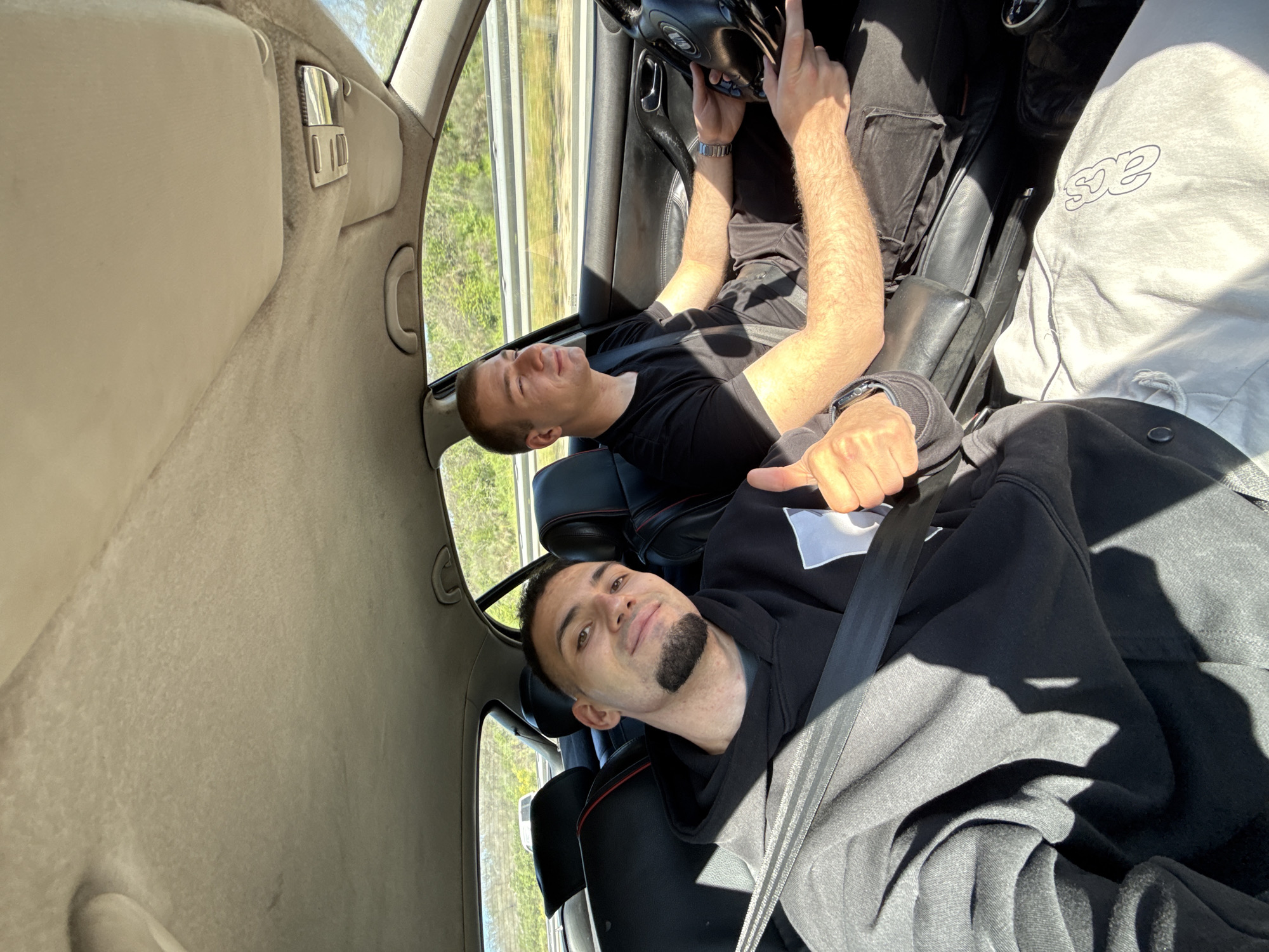 Two men are in the front seats of a car. The man in the passenger seat is driving. The man in the driver seat is turned towards the camera, giving a thumbs up. The location is not discernible.