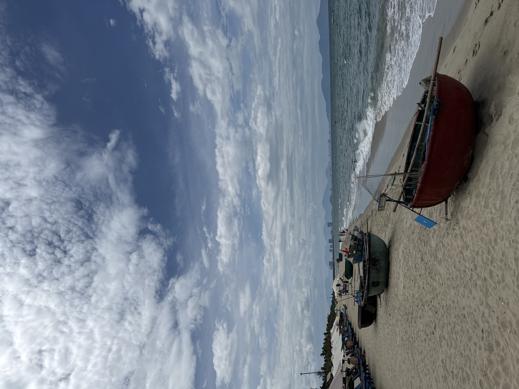 This is a medium shot of a beach in Hoi An, Vietnam. The sky is partly cloudy with a blue sky peeking through. The ocean is visible with gentle waves breaking on the shore. There are several traditional Vietnamese basket boats on the sand, with one red boat prominently in the foreground. Further down the beach, there are rows of lounge chairs with umbrellas, and a few people can be seen relaxing. In the distance, a city skyline with buildings and mountains can be observed. The sand is light brown and appears fine-grained.