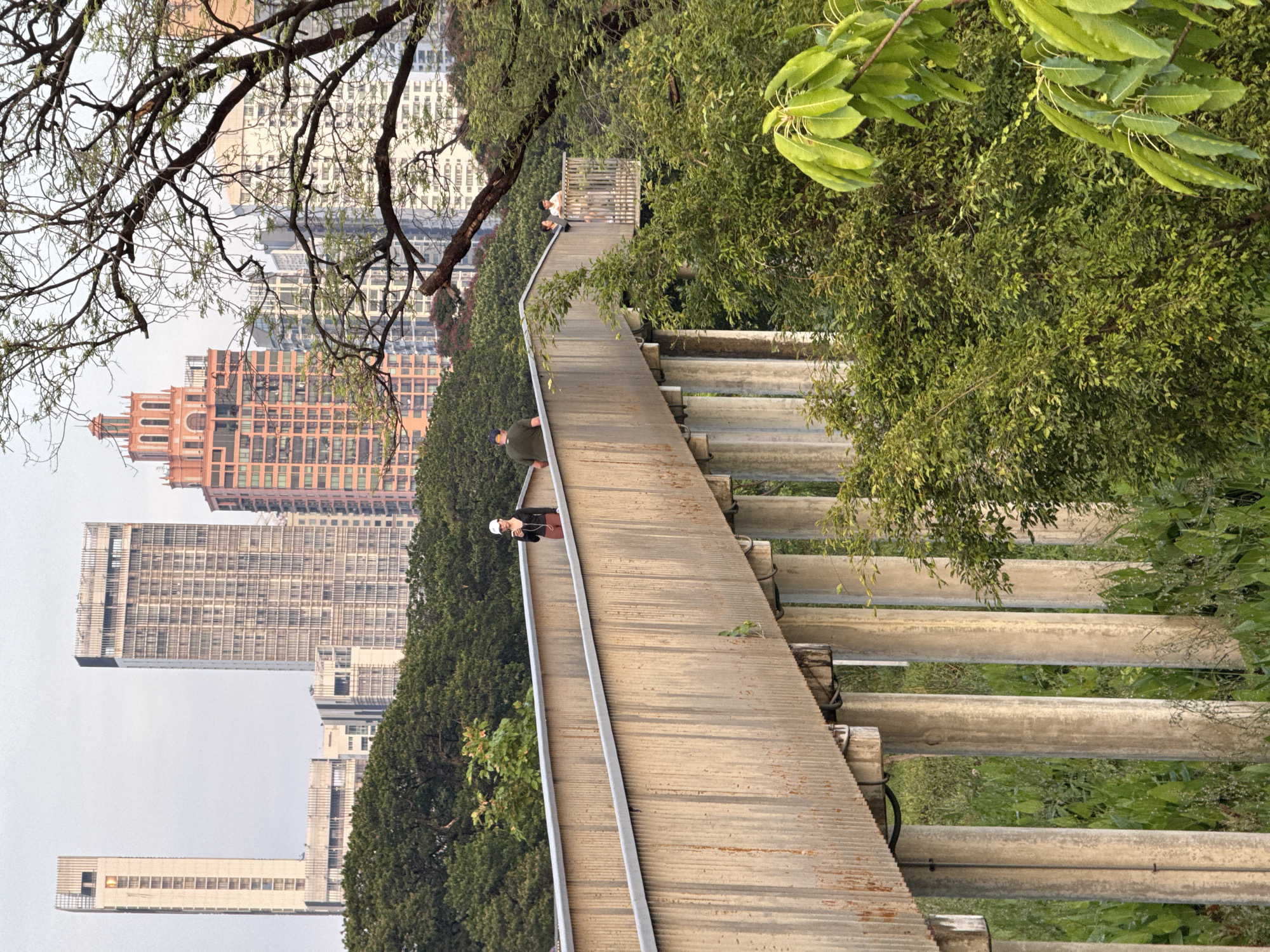 The image shows a view of the Bangkok Green Bridge, an elevated park built on a former railway track in Bangkok, Thailand. The bridge features a narrow walkway with metal railings and a series of concrete supports below. The walkway is elevated above the surrounding greenery. Several people are on the bridge, some walking, one in a white hat. In the background, there are modern skyscrapers. The surrounding environment includes trees and other vegetation. The sky is overcast.