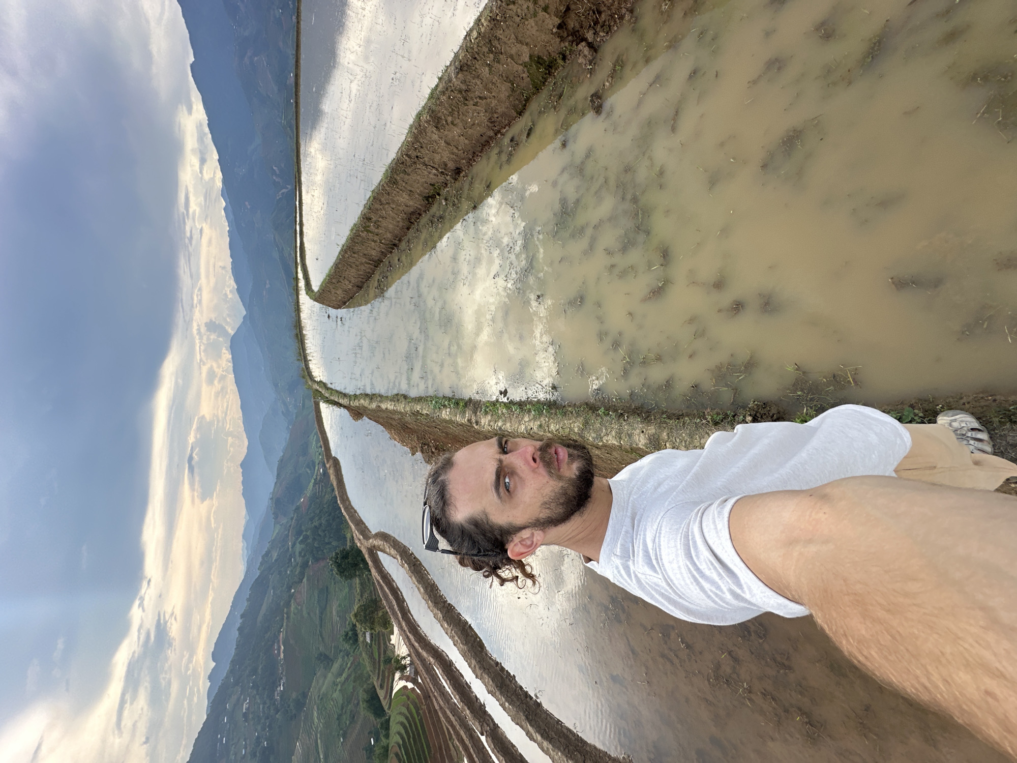 A man with a beard and curly dark hair, wearing a white t-shirt and sunglasses perched on his head, takes a selfie outdoors. He looks directly at the camera with a slightly pouting expression. His arm is extended, holding the camera, and part of his leg is visible.
The scene is dominated by terraced rice paddies, filled with water that reflects the cloudy sky. The paddies are arranged in curvilinear patterns, following the contours of the landscape, and separated by narrow earthen banks. Some visible plant matter is in the muddy water of the paddies. In the distant background, majestic mountains rise, layered under a partly cloudy sky with patches of bright white and darker grey clouds, suggesting either early morning or late afternoon light. The sky transitions from lighter hues near the horizon to a deeper blue overhead. The overall impression is one of a rural, mountainous agricultural landscape. There is no visible text.