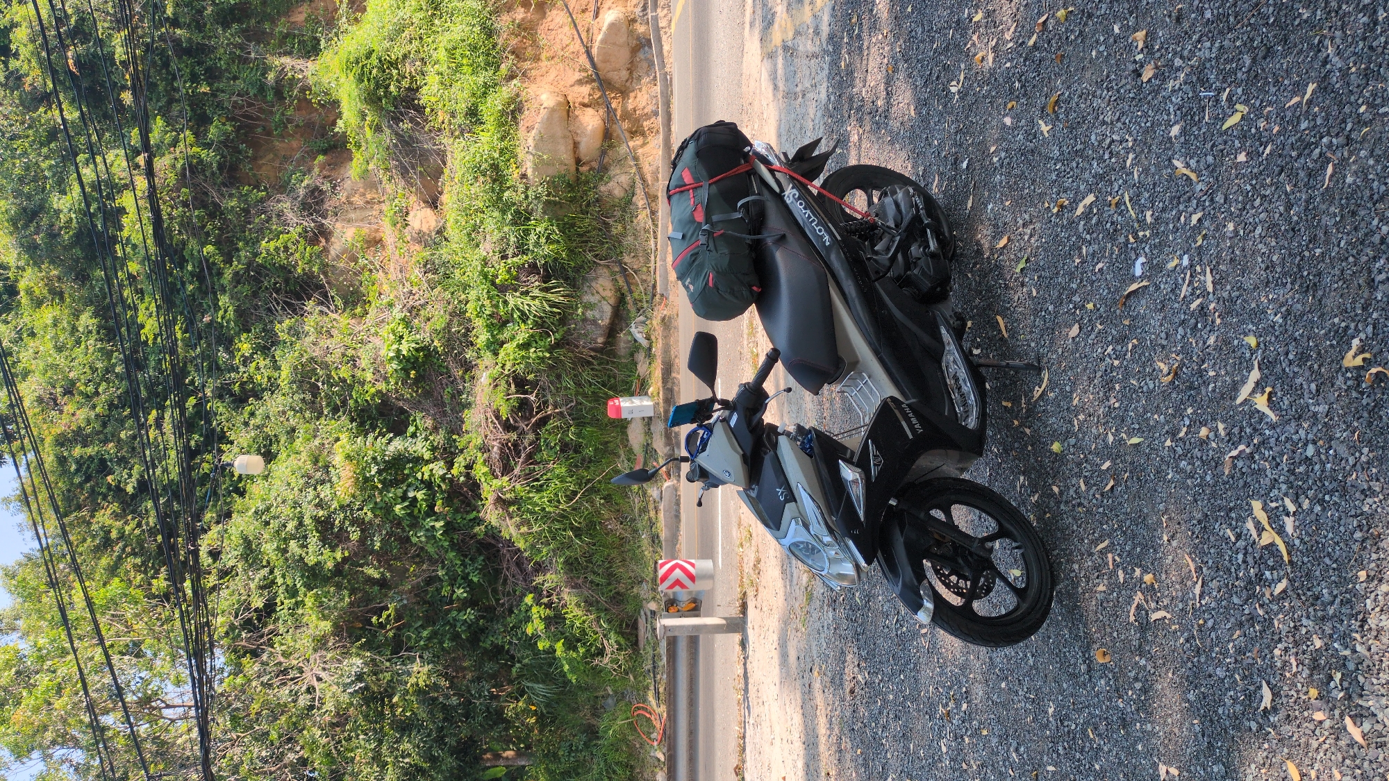 The image shows a Yamaha motorcycle parked on the side of a road in Đại Lãnh, Vietnam. The road is lined with greenery and trees on one side. A bag is strapped to the back of the motorcycle. A roadside marker and guardrail are visible near the motorcycle. The ground is covered with gravel.