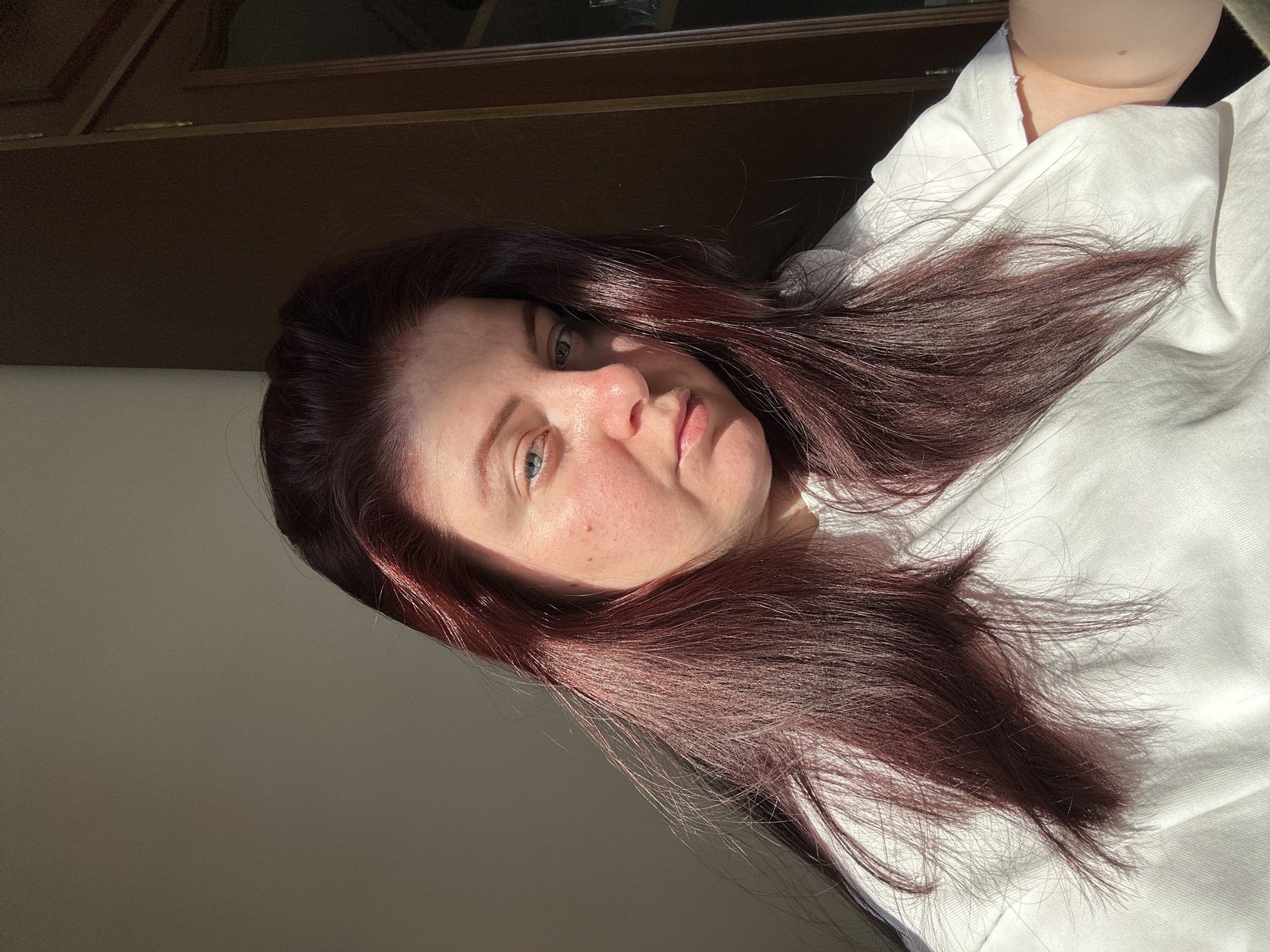 A woman with long, dark red hair is positioned in the foreground of the image, looking towards the camera. She is wearing a white t-shirt. The lighting suggests an indoor setting with natural light casting shadows on her face and hair. The background is divided, with a plain, light-colored wall on the left and a dark wooden surface, possibly a door or cabinet, on the right. No specific landmarks of the city of Rijeka, Croatia are visible.