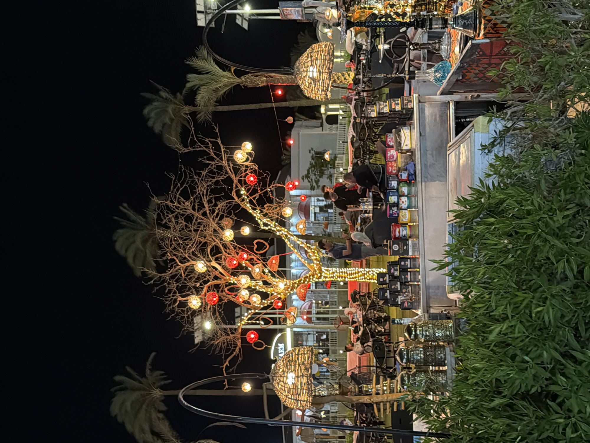 This is a night-time photograph of an outdoor seating area at a cafe or restaurant in Sharm El Sheikh, Egypt. The scene is decorated with numerous lights, including string lights and lanterns hanging from trees and poles. In the foreground, there is lush green foliage. Behind the foliage, tables and chairs are set up for patrons, some of whom are seated and appear to be dining or socializing. A prominent element is a bare tree adorned with many colorful, glowing spherical lanterns and heart-shaped ornaments. Several people are visible in the mid-ground, some standing and interacting, while others are seated at tables. In the background, there are buildings and palm trees, characteristic of the Egyptian coastal city. The overall atmosphere is festive and inviting, illuminated by the warm glow of the decorative lighting.