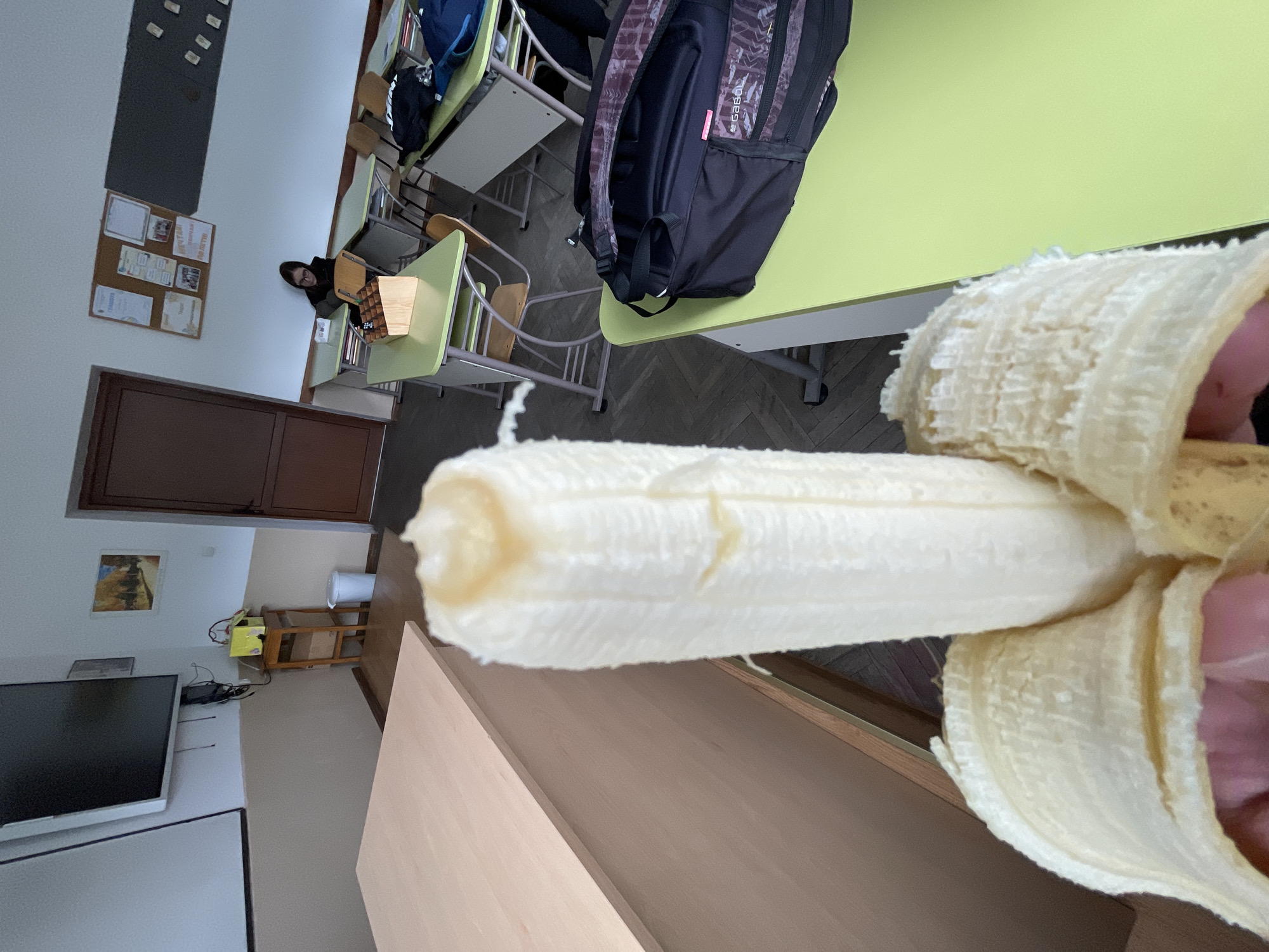 The image shows a peeled banana held in the lower right foreground, with its peel and the hand holding it visible. The setting appears to be a classroom, with desks arranged in rows and a person sitting at one of them in the background. A backpack is on the desk closest to the banana. In the background to the left, there's a wall-mounted TV and a door. There is no information provided in the image to confirm that this location is the city of Zlatograd, Bulgaria.