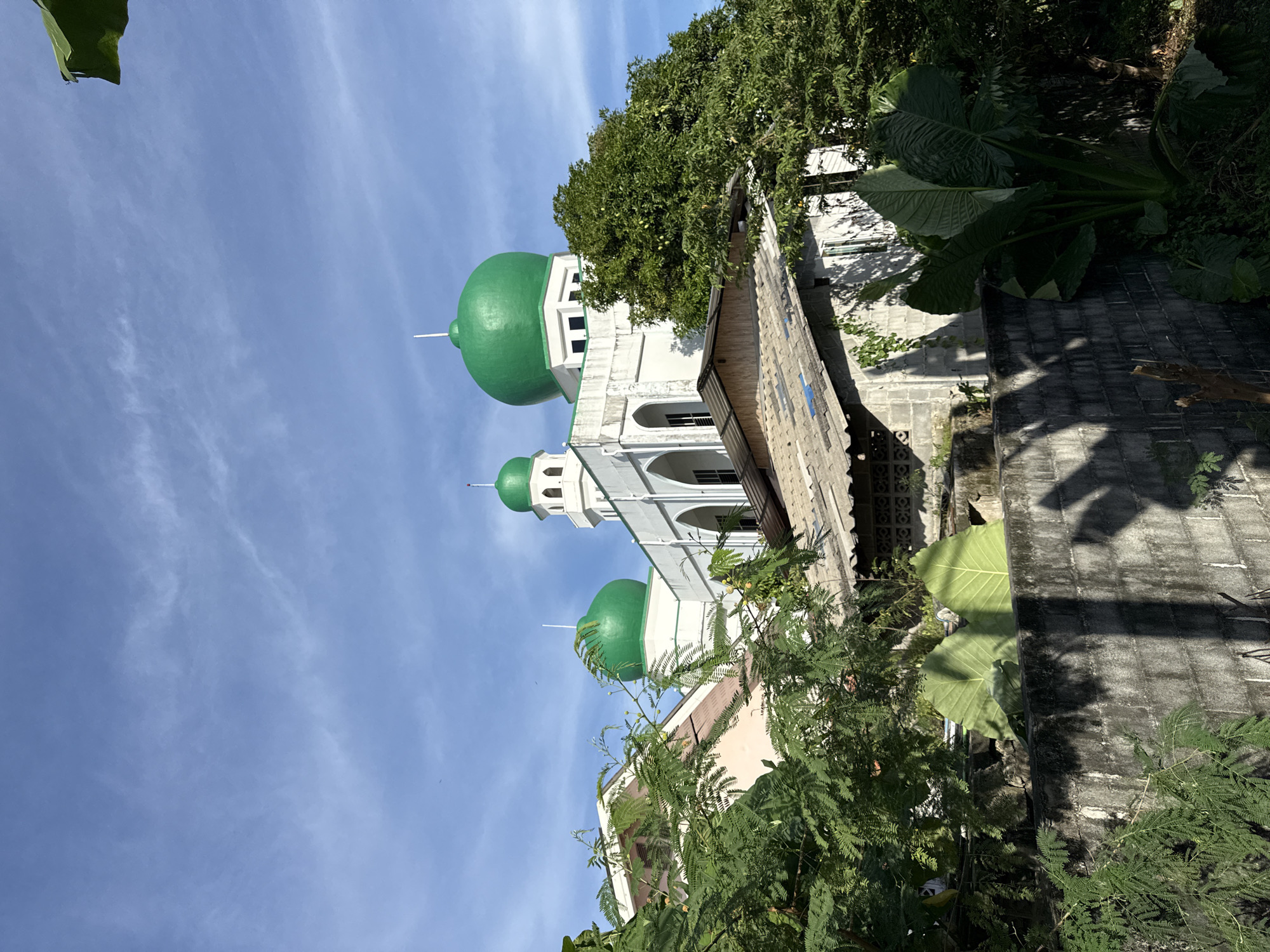 The image displays a mosque with green domes, a minaret, and white architecture against a bright blue sky with scattered clouds. Lush green trees and foliage surround the building. In the foreground, a grey concrete wall casts shadows. The scene is likely situated in Phuket City Municipality, Thailand, given the architectural style and the prevalent vegetation. There are no people visible in the image.