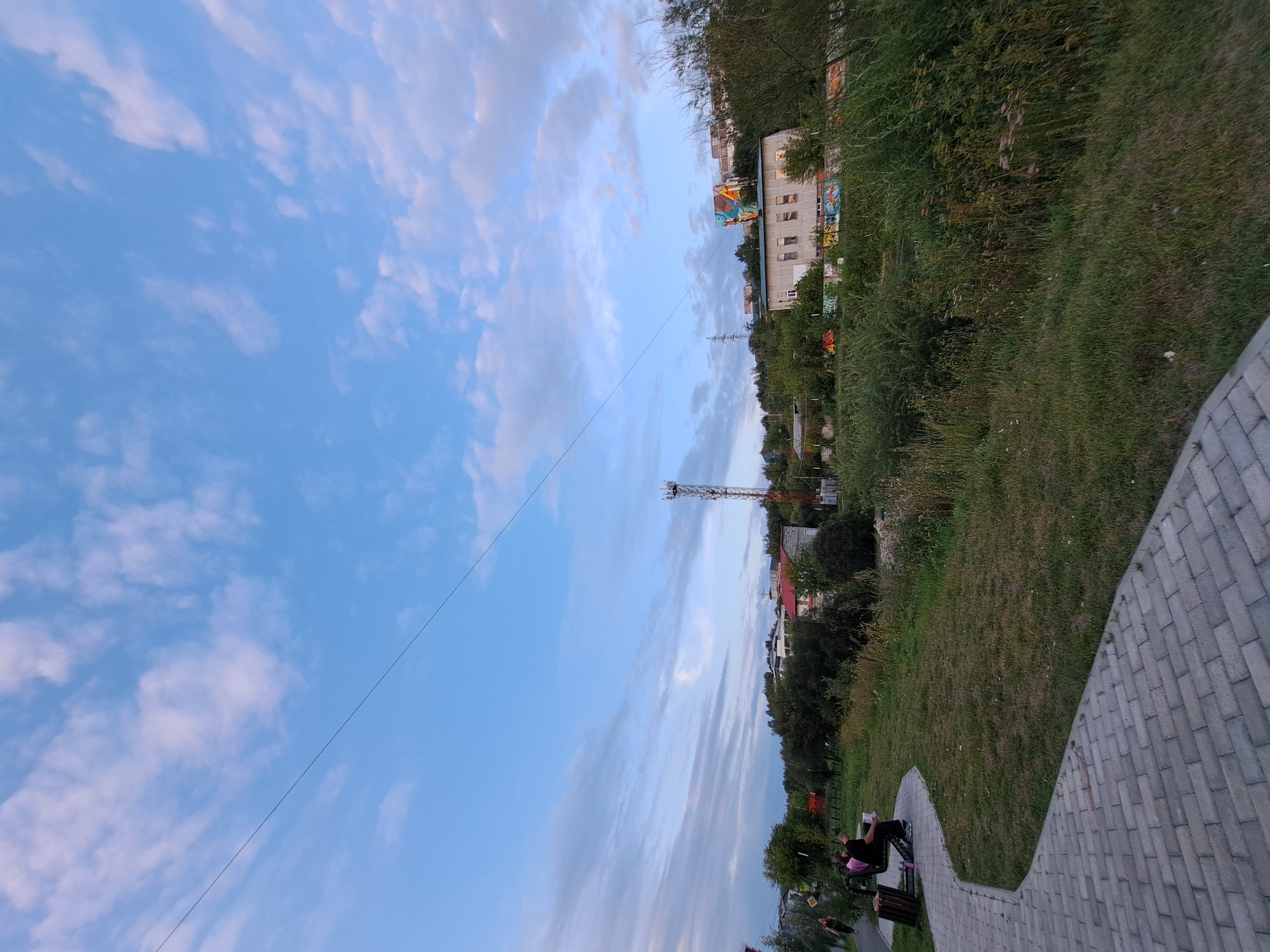The image depicts a scene in a park area, possibly on the outskirts of a city, featuring a paved walkway, grassy areas, and trees. In the background, there are several buildings, a telecommunications tower, and a structure with colorful graffiti or artwork. The sky is partly cloudy, with the sun appearing to be setting or rising, casting a soft light on the scene. Two individuals are seated on a bench on the left side of the image, facing away from the camera. The overall atmosphere is calm and serene. The image appears to be taken in the city of Irkutsk, Russia.
