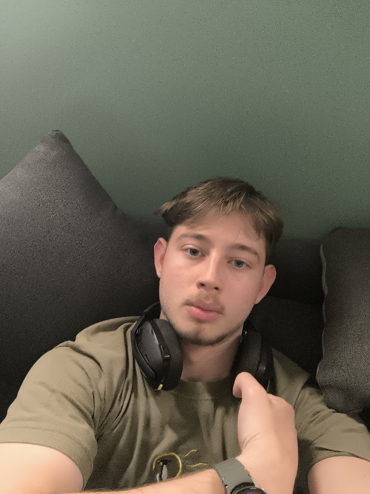 A medium close-up shot shows a young Caucasian male. He has short brown hair and blue eyes, and a light beard. He is wearing a dark olive green T-shirt and black headphones around his neck. He is wearing a grey smartwatch on his left wrist and resting his left hand on his chest. He is sitting on a dark grey, cushioned sofa in front of a solid green wall. The photo does not contain any information indicating location or that it was taken in Sofia, Bulgaria.