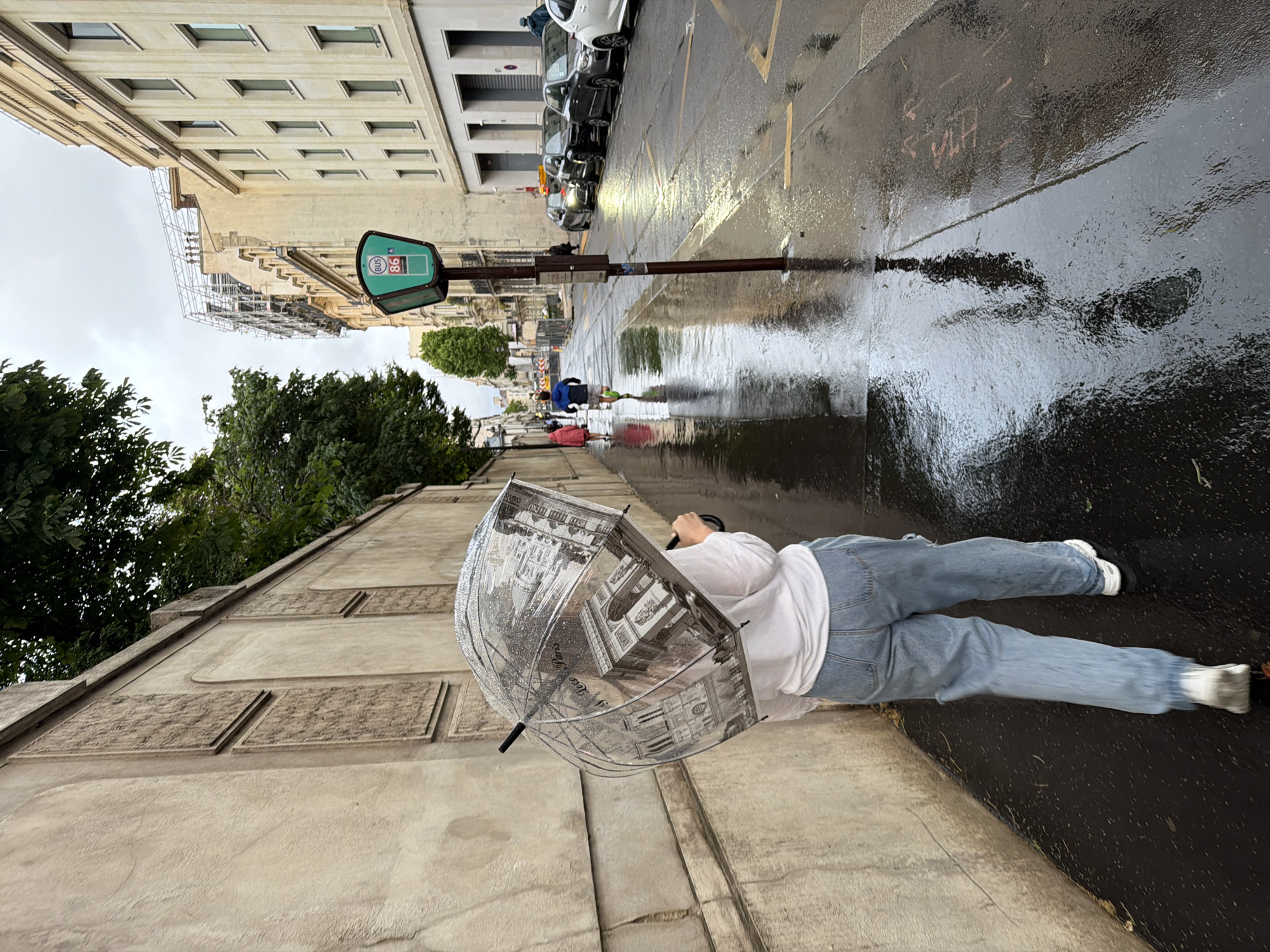 The image depicts an urban street scene on an overcast day, likely after or during rain, given the wet, reflective asphalt and the use of an umbrella. The perspective is from a slightly elevated position, looking down the street.
In the foreground, a person is walking or standing on a concrete sidewalk, holding a clear umbrella. The person is wearing a white t-shirt, blue jeans, and white sneakers. The umbrella features black and white illustrations of Parisian landmarks such as the Arc de Triomphe, the Eiffel Tower, and Sacré-Cœur, with visible text "Paris" and "Je t'aime" printed on it, strongly indicating the location is Paris, France.
To the left of the street, there's a multi-story building with numerous windows, and scaffolding is visible on a section of it. Lush green trees line the sidewalk further down the street. On the right side of the street, a row of parked cars, predominantly dark-colored, is visible next to another building.
A green bus stop sign, typical of Paris, stands prominently on a pole in the middle ground, displaying "BUS" along with route numbers "86" and "98". The wet street reflects the surrounding buildings, cars, and sky, creating shimmering puddles and distorted reflections. Faint reddish graffiti, possibly "FHH", and an arrow can be seen on the wet ground. The overall atmosphere is that of a cool, possibly damp, urban day.
