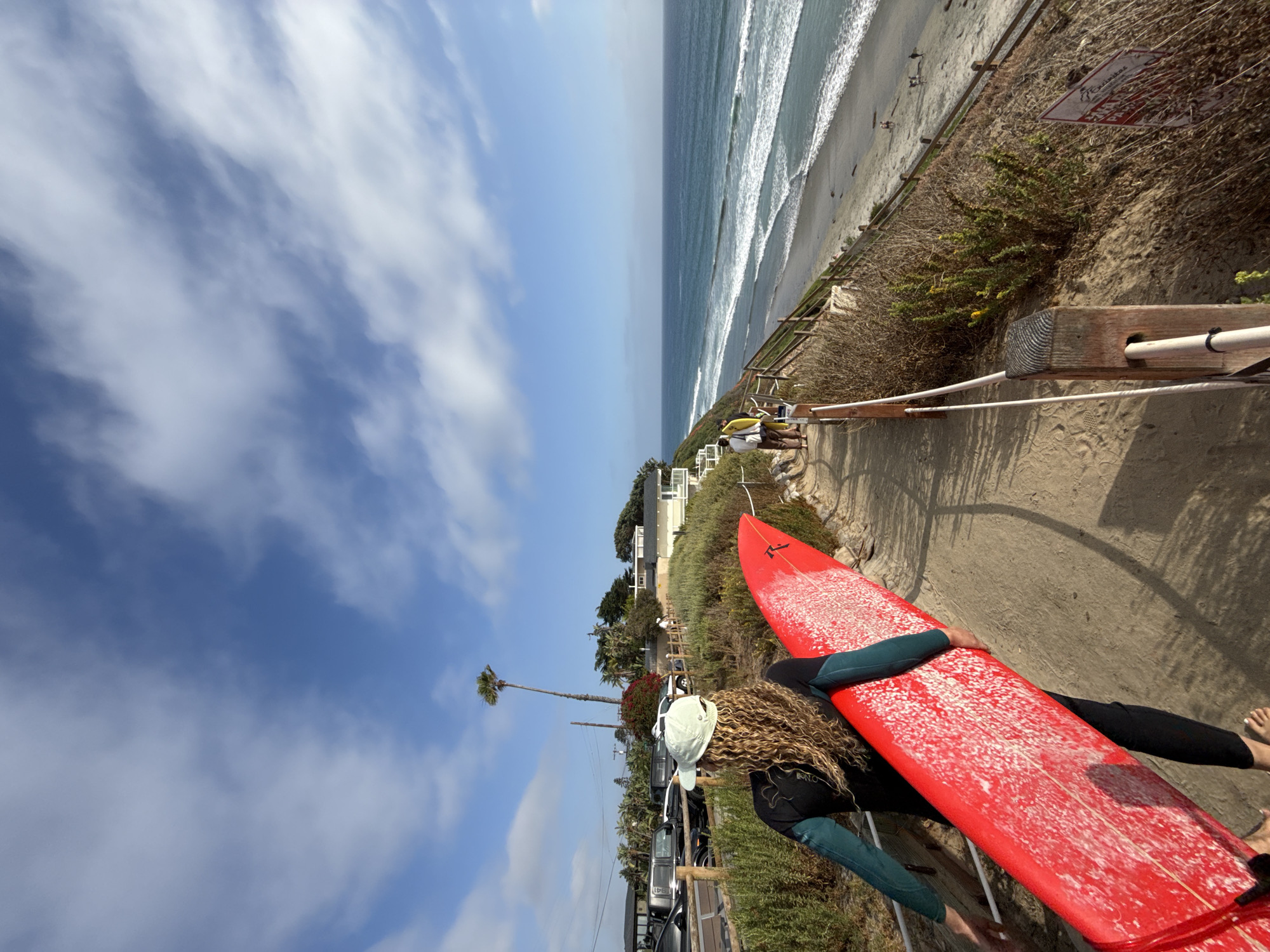 The image depicts a bright, sunny coastal scene in Encinitas, United States, viewed from a slightly elevated sandy path. In the foreground, a person with long, curly light brown hair, wearing a dark wetsuit with "O'NEILL" visible on the sleeve and a light-colored cap, carries a vibrant red surfboard. The surfboard, which has "RUSTY" visible on its surface, is covered in white wax marks.
The path the person is on runs along a sandy cliffside, bordered by wooden posts and white ropes acting as a railing. Beyond this railing, coastal vegetation grows on the slope. Further down the cliff, a red sign with white text is visible, partially obscured, which appears to be a warning or informational sign, possibly mentioning "DANGER".
In the midground and background, the Pacific Ocean stretches out under a clear blue sky dotted with scattered white clouds. Waves break along the sandy shoreline, where a few distant figures can be seen. On the land side, beyond the path, several houses with white and light-colored facades are visible, nestled amidst green trees, including tall palm trees. Parked vehicles are also discernible along what appears to be a street or driveway. The scene suggests a pleasant day for outdoor activities like surfing, with the sun casting clear shadows.