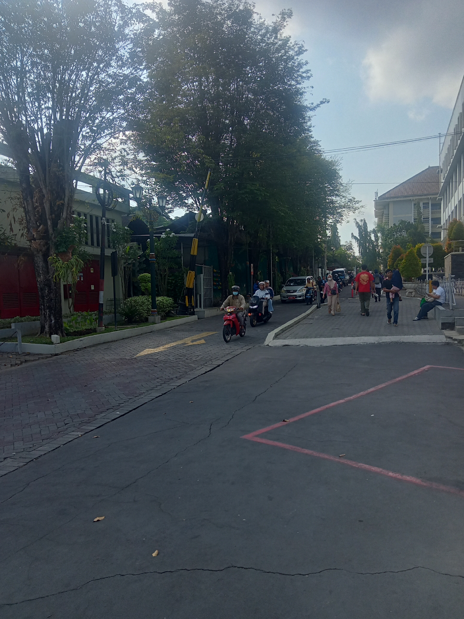 The image depicts a street scene in Depok, Indonesia, likely on a university campus given the presence of students and faculty. On the left side of the frame, a brick-paved road leads towards a building with red doors and manicured greenery. A yellow arrow painted on the road indicates a direction of travel. Several people are walking and riding motorcycles along the street. In the foreground, a dark asphalt road shows some cracks and fallen leaves. In the background, a modern building with white vertical elements and a tiled roof is visible. The sky is partly cloudy. The overall impression is of an active, urban environment with a mix of natural and architectural elements.