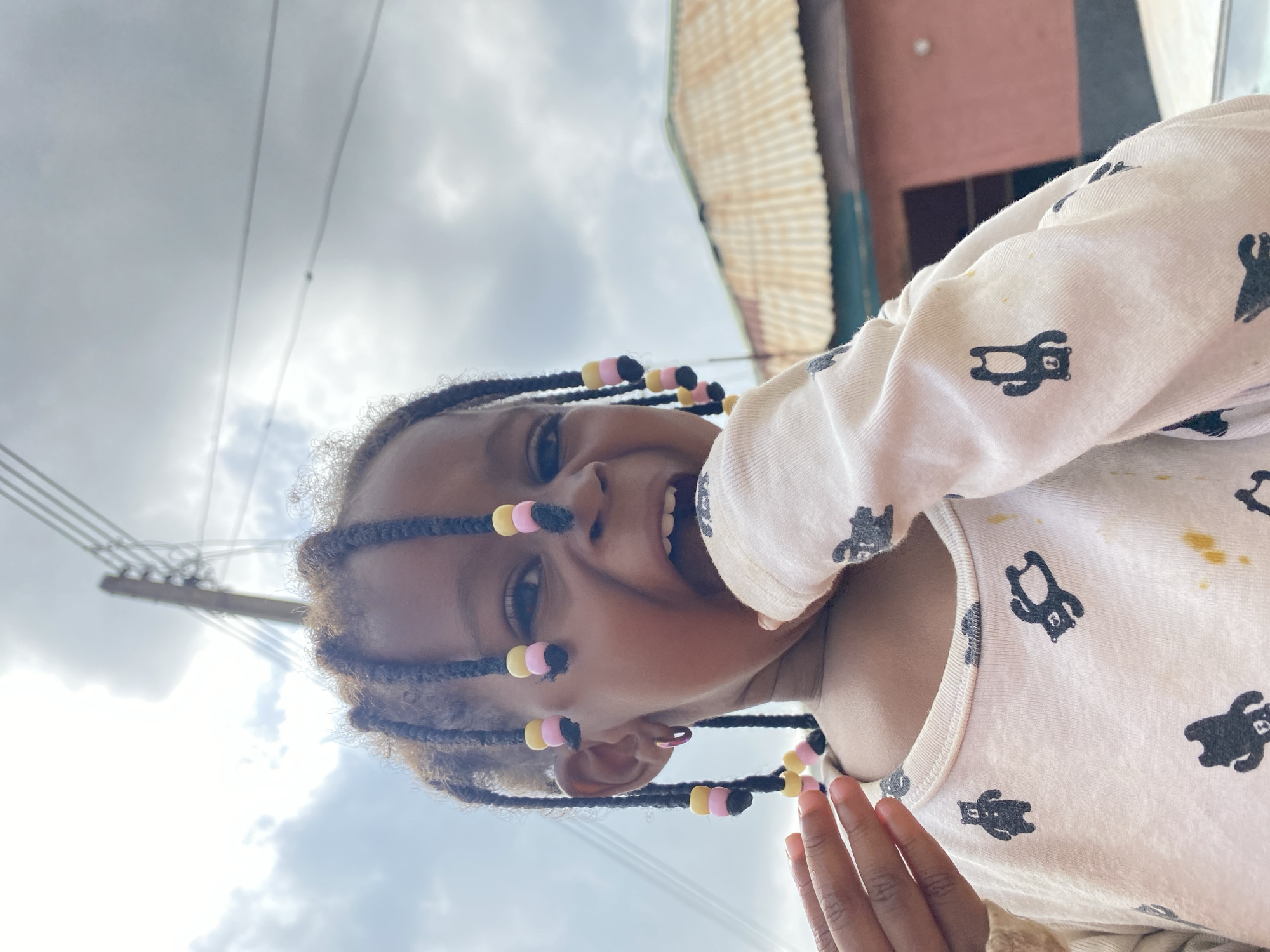 A close-up shot captures a young Black child with braided hair adorned with colorful beads, looking directly at the camera with a playful expression. The child's mouth is open, and their hand is near their face. They are wearing a light-colored long-sleeved shirt with a pattern of dark animal silhouettes. In the background, a corrugated metal roof of a building is visible, along with a utility pole and overcast sky. The setting appears to be an outdoor urban environment, consistent with the city of Bukuru, Nigeria.