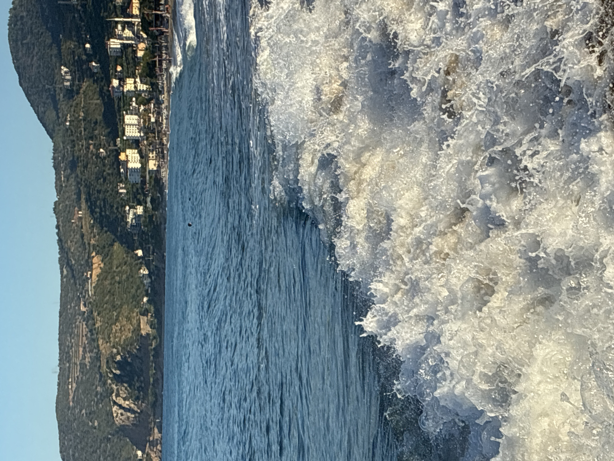 The image shows a close-up view of a crashing wave on a beach, with a town nestled on a hillside in the background. The foreground is dominated by white foam and spray from the breaking wave, which obscures the lower part of the scene. The water is a vibrant blue, with smaller ripples visible beyond the main wave. In the mid-ground, a strip of sandy beach is visible, dotted with people and umbrellas. Behind the beach, buildings of various sizes and colors are clustered on the slopes of a large, green hill. The architecture suggests a Mediterranean coastal town, and the presence of a minaret indicates a location with a significant Muslim population, consistent with Alanya, Turkey. The sky is a clear, pale blue, indicating a sunny day. The overall impression is of a lively beach scene during daylight.