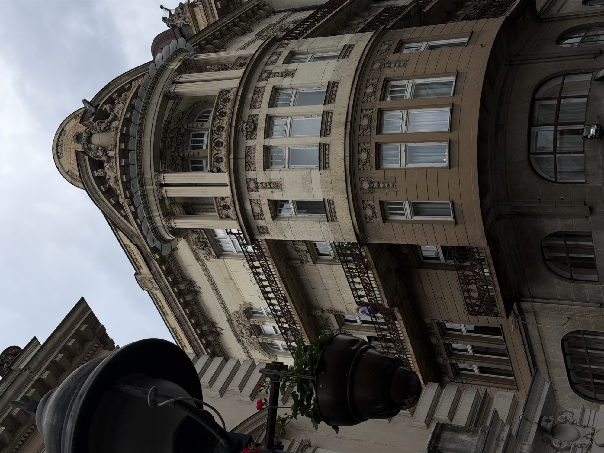 A low-angle shot depicts a multi-story building in Belgrade, Serbia, characterized by its curved facade and ornate architectural details. The building's color scheme transitions from lighter shades on the upper floors to darker tones below. Windows punctuate the facade, some with balconies featuring wrought-iron railings. A street lamp in the foreground, partially obscured by a flower pot with greenery and red blossoms, adds depth to the composition. The overcast sky provides soft lighting, emphasizing the building's texture. A figure in a light-colored shirt is visible on one of the balconies.