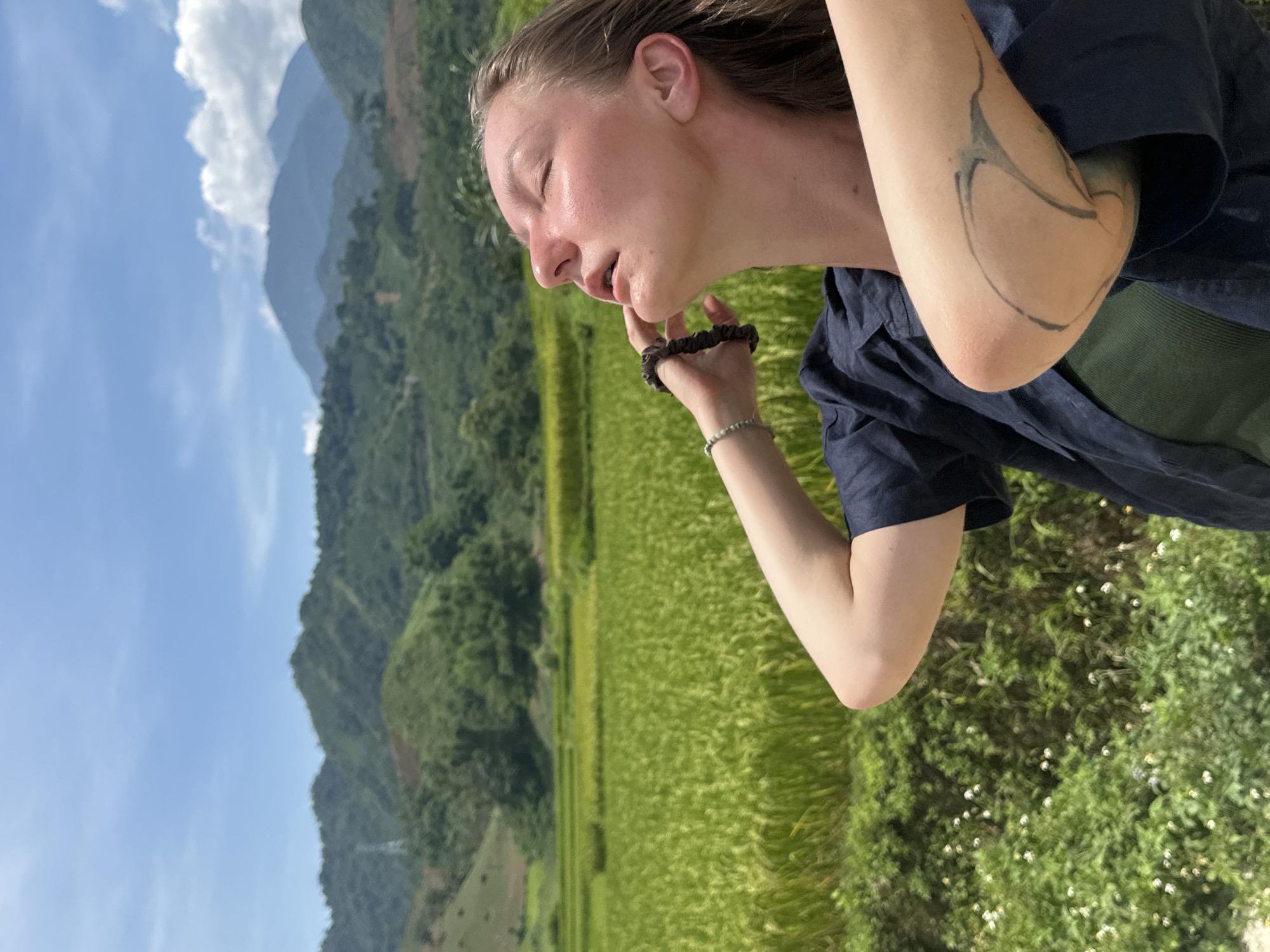 This portrait-oriented image captures a person outdoors against a scenic natural backdrop. The central subject is a young adult with fair skin and light brown hair, positioned on the right side of the frame. Their eyes are closed, and their head is tilted back, with strong sunlight illuminating their face, giving it a somewhat flushed appearance. Their lips are slightly parted. One hand is raised to their face, fingers gently near their chin, wearing a dark elastic hair tie or scrunchie and a light-colored beaded bracelet on the wrist. They are dressed in a dark blue collared shirt worn open over a dark green undershirt. An abstract tattoo with black lines is visible on their right forearm/elbow area.
The background features expansive, vibrant green fields, typical of rice paddies, stretching across the mid-ground. Beyond the fields, majestic mountains rise into the distance, covered in lush greenery. The sky above is clear and blue with scattered white clouds, indicating bright daytime. In the immediate foreground, to the bottom right, are patches of green foliage with small white flowers.
The overall impression is one of peaceful contemplation or enjoyment of the natural environment, possibly feeling the warmth of the sun. The setting strongly suggests a rural, mountainous, and agricultural region, consistent with the provided context of Duong Quy, Vietnam. There is no visible text in the image.