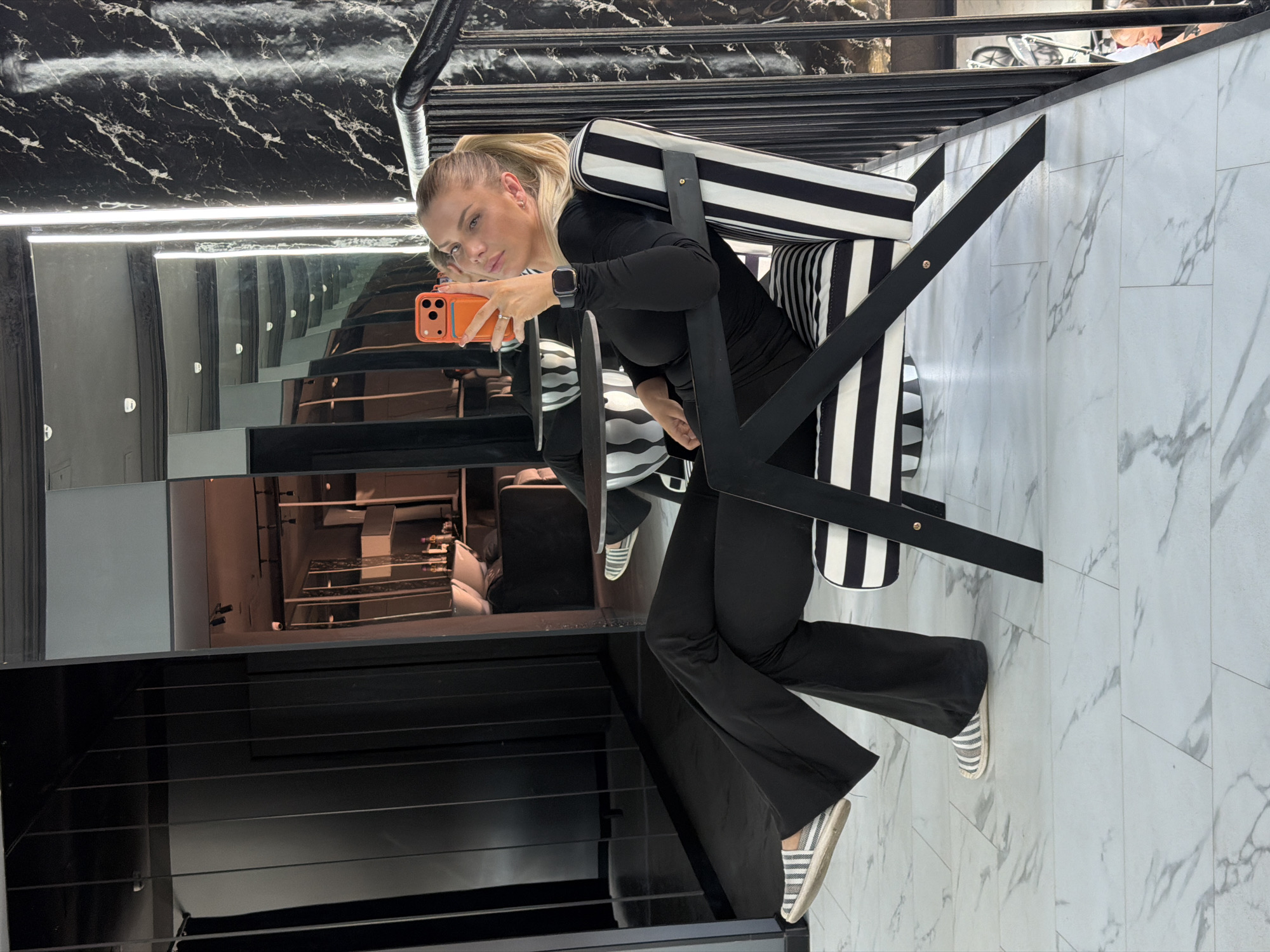 The image shows a blonde woman in her late twenties or early thirties taking a selfie in a modern interior. She is wearing a black long-sleeved top and black flared pants. She is sitting in a chair with a black and white striped cushion and a black frame. Her right leg is crossed over her left. She is holding an orange smartphone in her right hand. The background appears to be a reflection in a large mirror. The reflection shows a contemporary bathroom or spa area with sinks, mirrors, and lighting fixtures. The overall aesthetic is very monochromatic and sleek, with black, white, and metallic accents. The flooring is white with grey marble-like veining. The image was taken in Moscow, Russia, at a location with a luxurious and modern design.