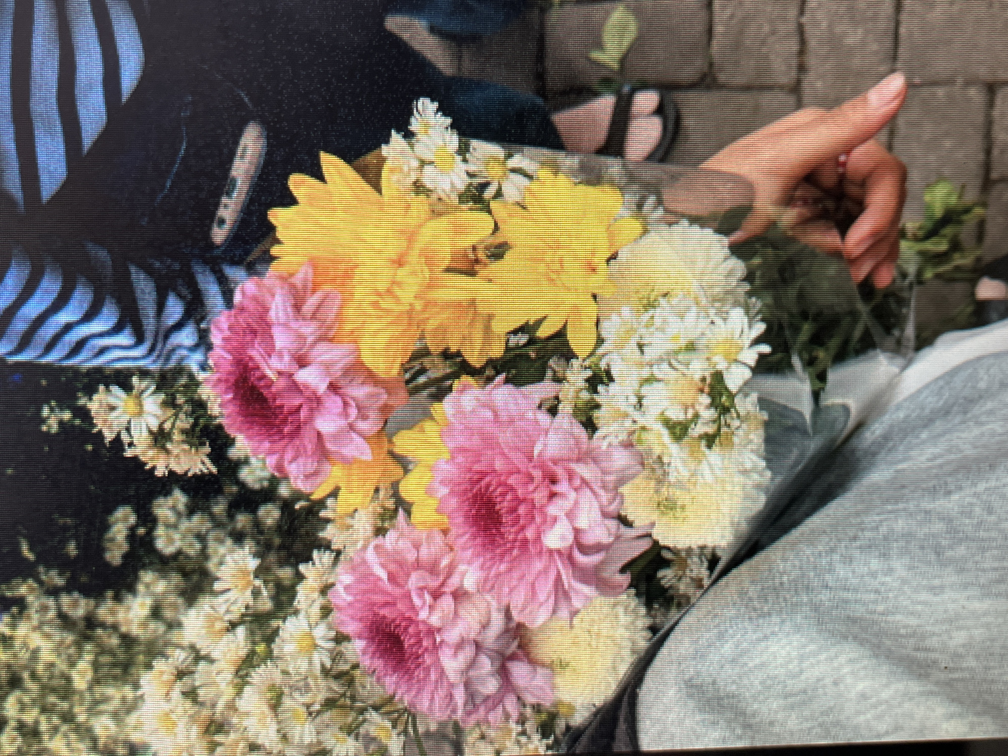 In Sejahtera, Indonesia, a close-up shot focuses on a bouquet of flowers, featuring large pink and yellow blooms interspersed with smaller white flowers. A person's hand, holding the bouquet, is visible on the right side of the frame, with fingers gently grasping the stems. The background shows parts of a person wearing a black garment with blue stripes and a black balaclava. The ground beneath appears to be paved with brick-like stones. The overall scene suggests an outdoor setting, possibly a street or garden in the city.
